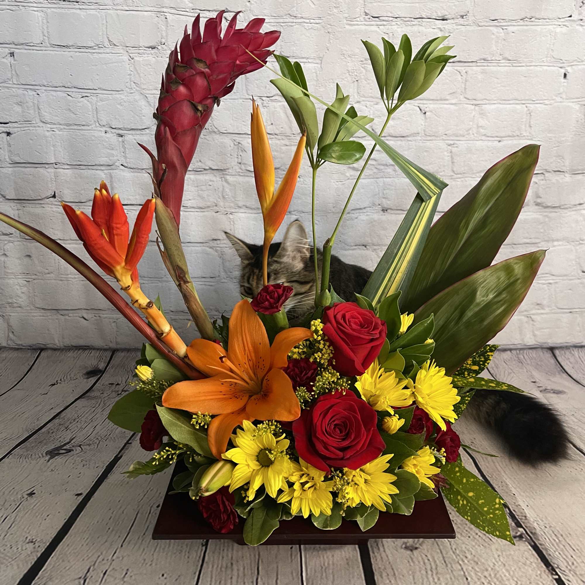 Tropical flower arrangement with red roses, orange lily, and yellow daisies in a low wooden base