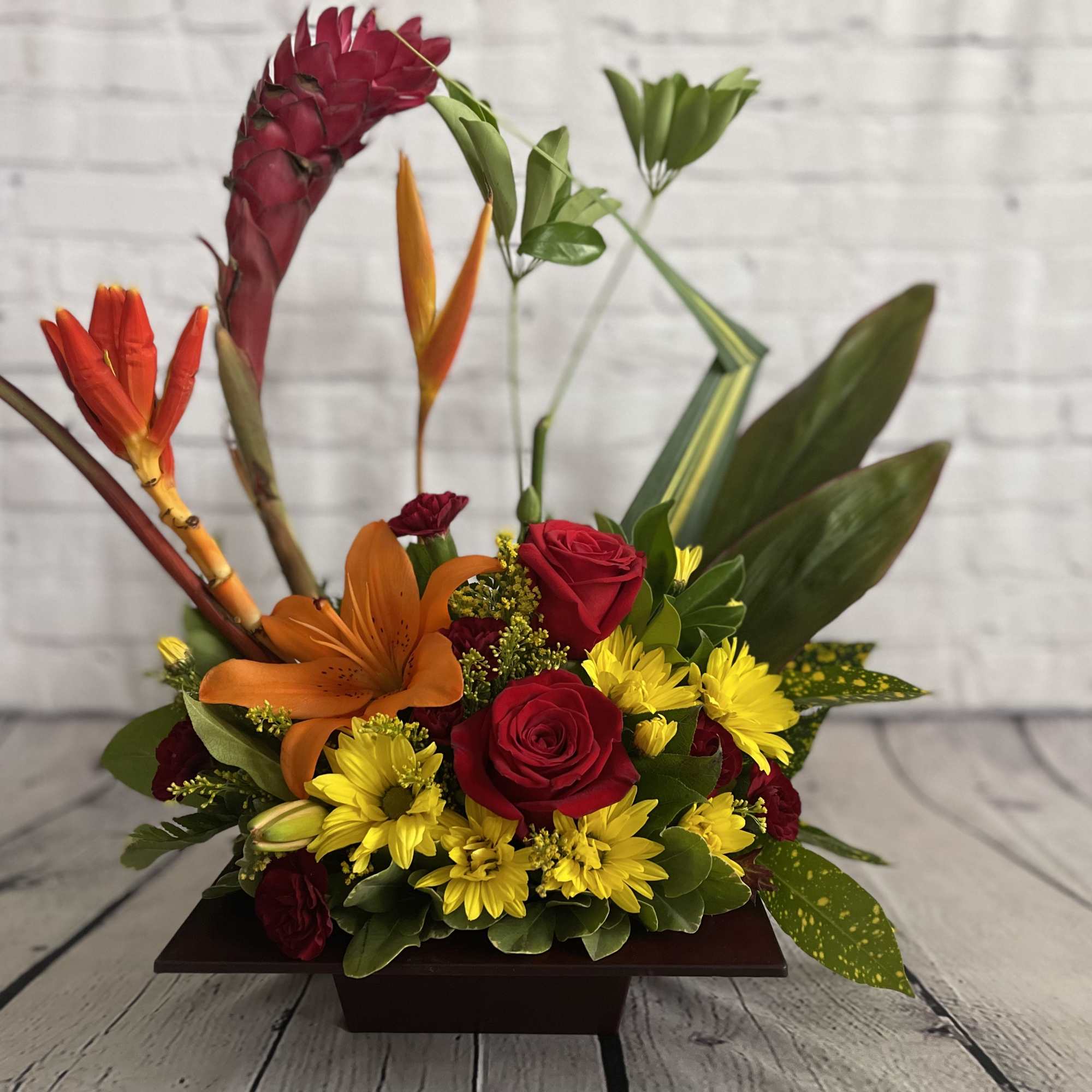 Tropical flower arrangement with red roses, orange lily, and yellow daisies in a low wooden container