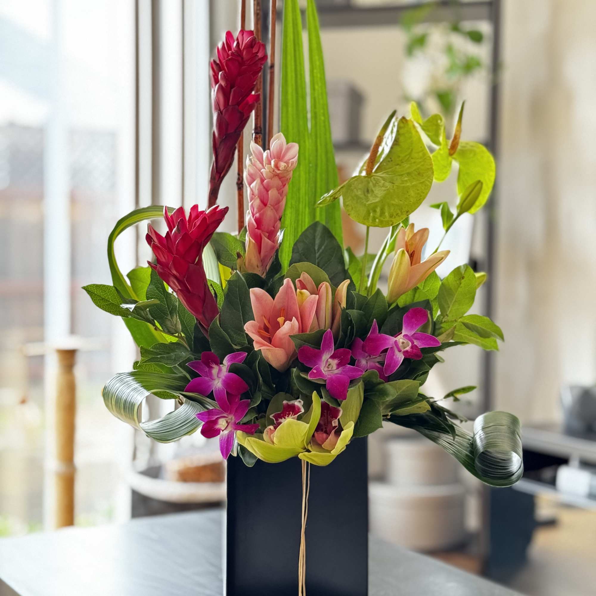Tall tropical arrangement with pink gingers, green anthuriums, and purple orchids in a black vase