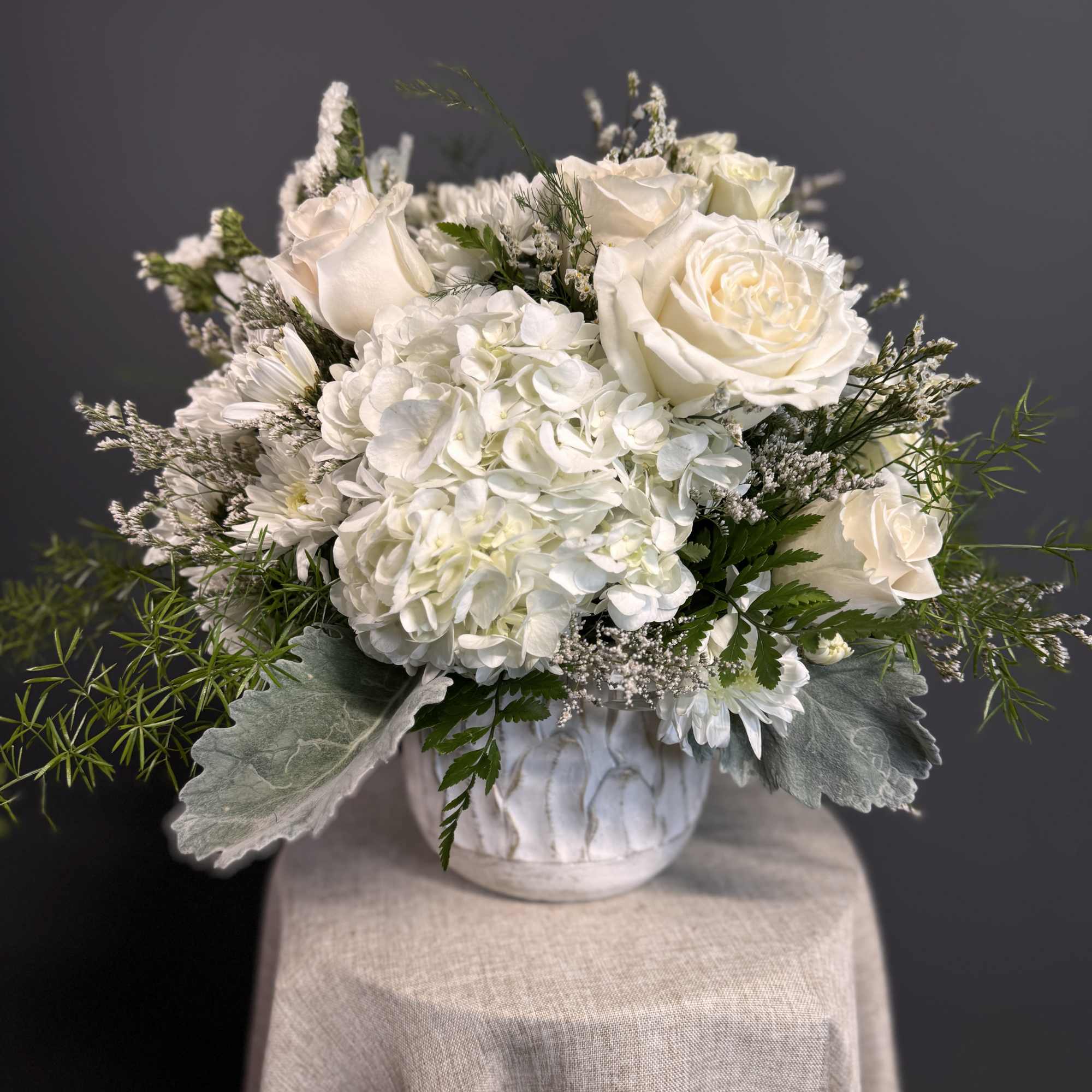 Low white arrangement of roses and hydrangeas in a white ceramic pot on a draped pedestal