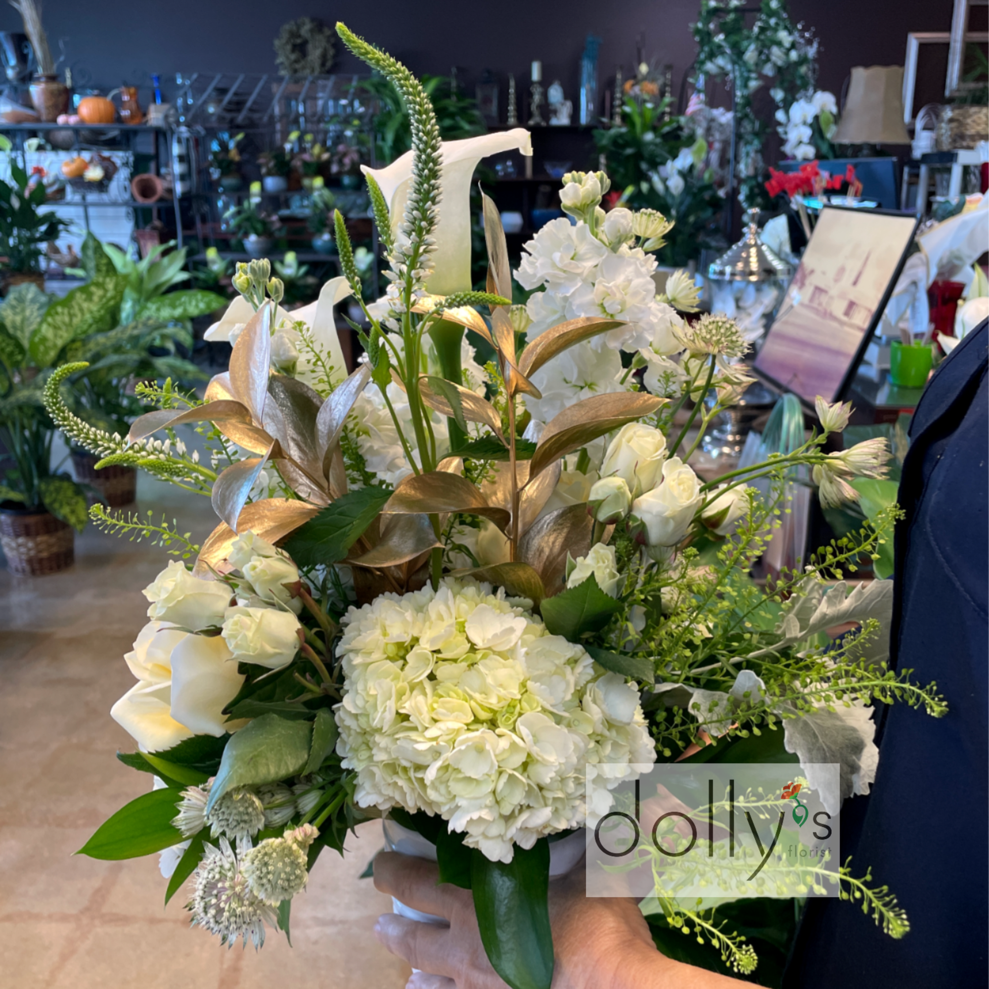 Compact white arrangement of hydrangeas, roses, and calla lilies in a small vase with gold accent leaves.
