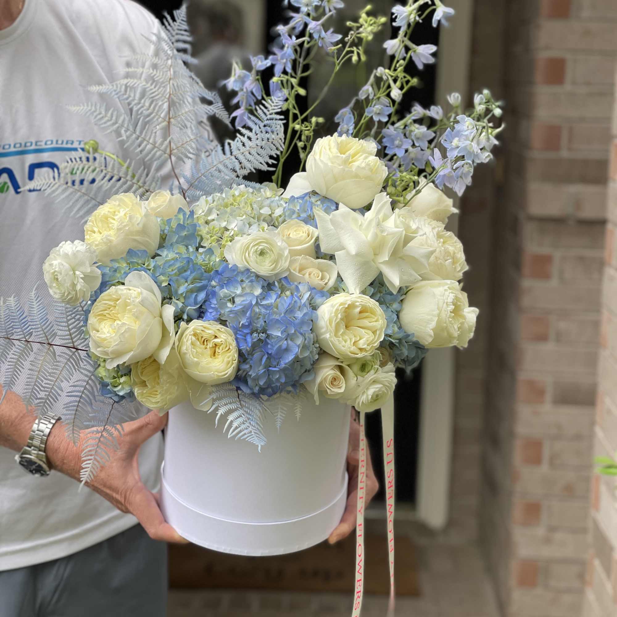 White hatbox arrangement with cream roses, blue hydrangeas, and pale blue delphinium