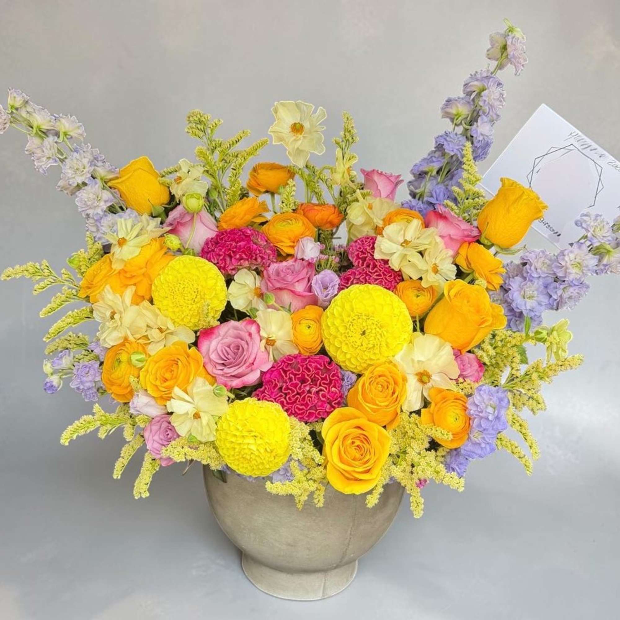 Bright mixed bouquet in a round vase with yellow, pink, orange, and lavender flowers.