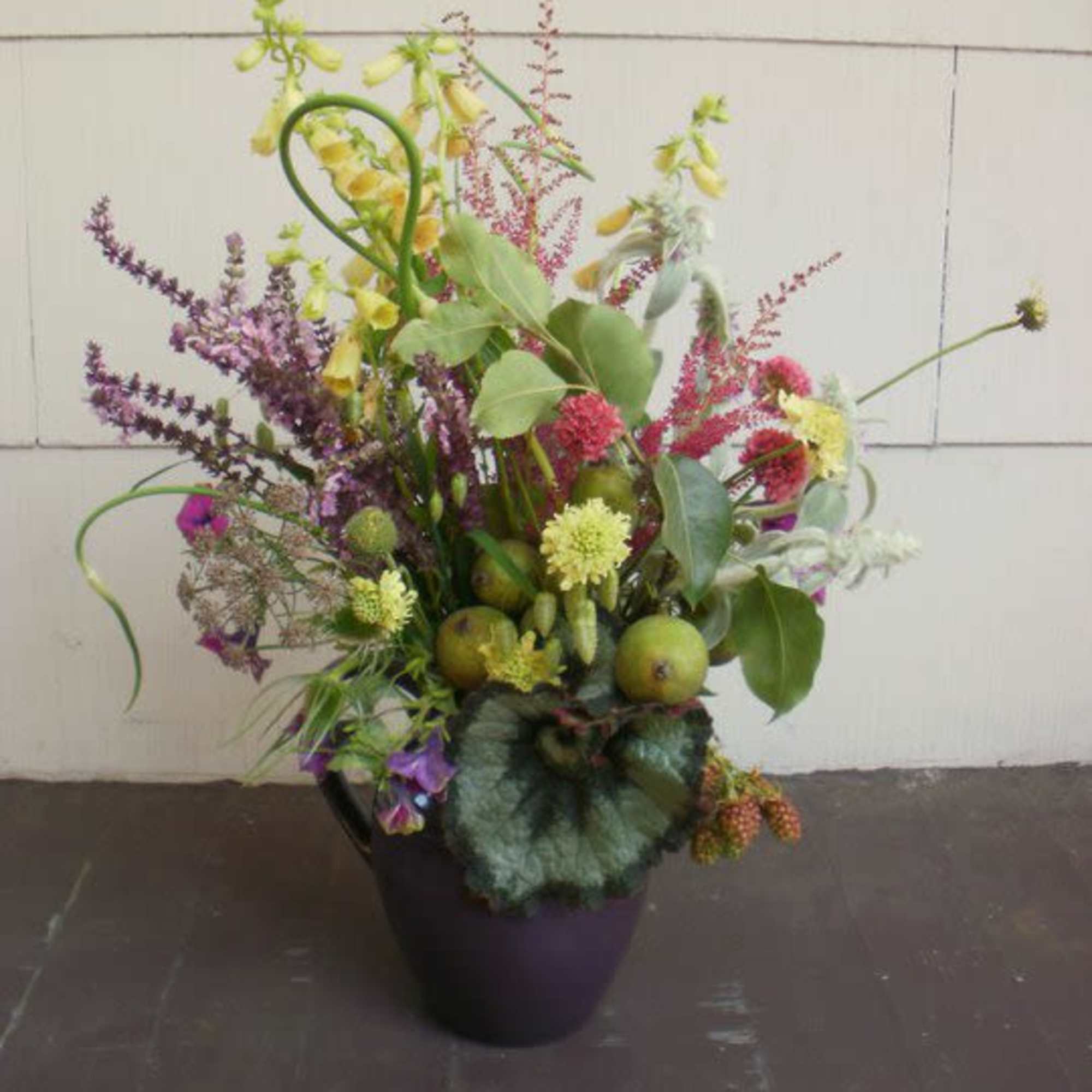 Wild-style arrangement of yellow and purple flowers with green fruits in a dark purple vase