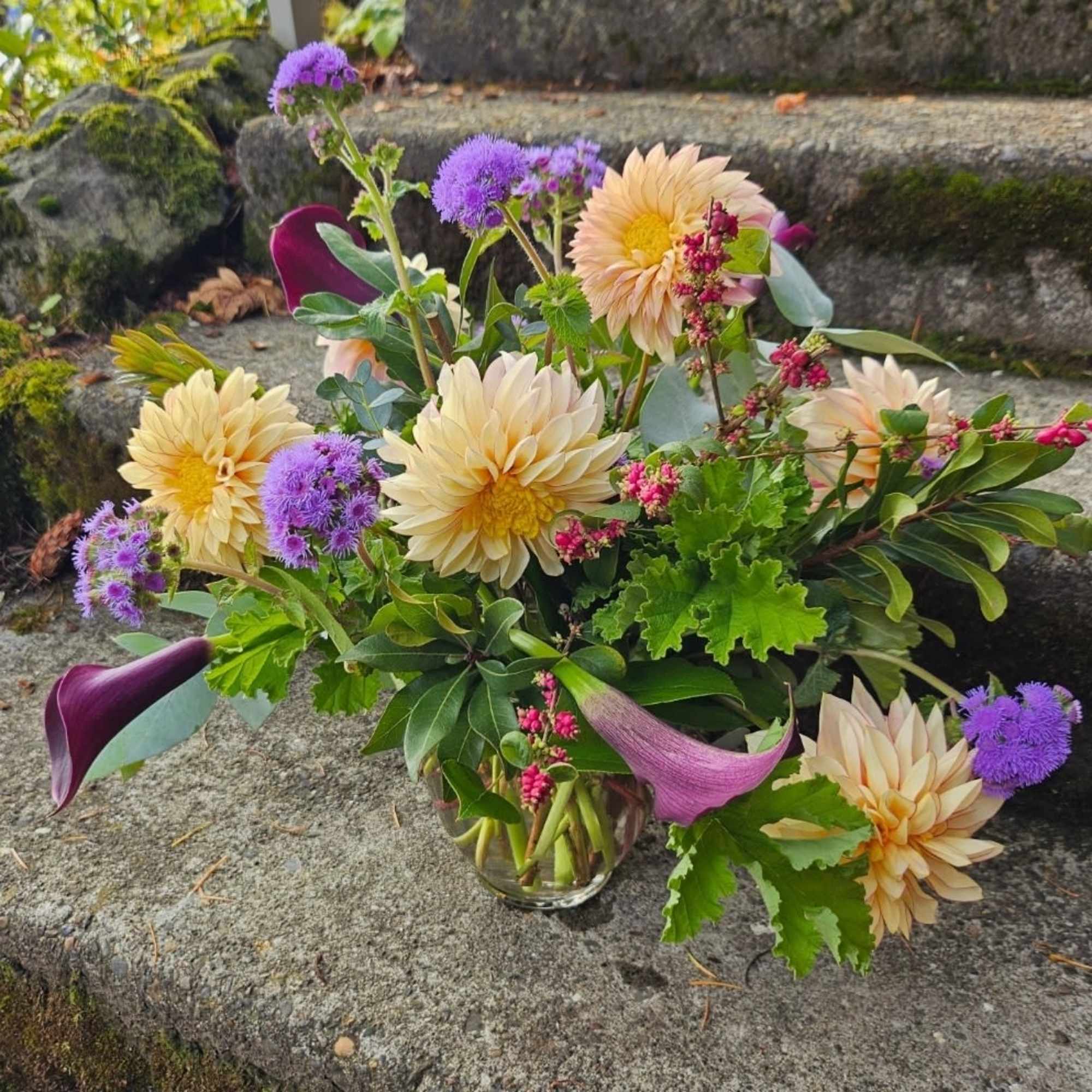 Low glass vase of peach dahlias, purple flowers, and dark plum calla lilies in a loose garden style