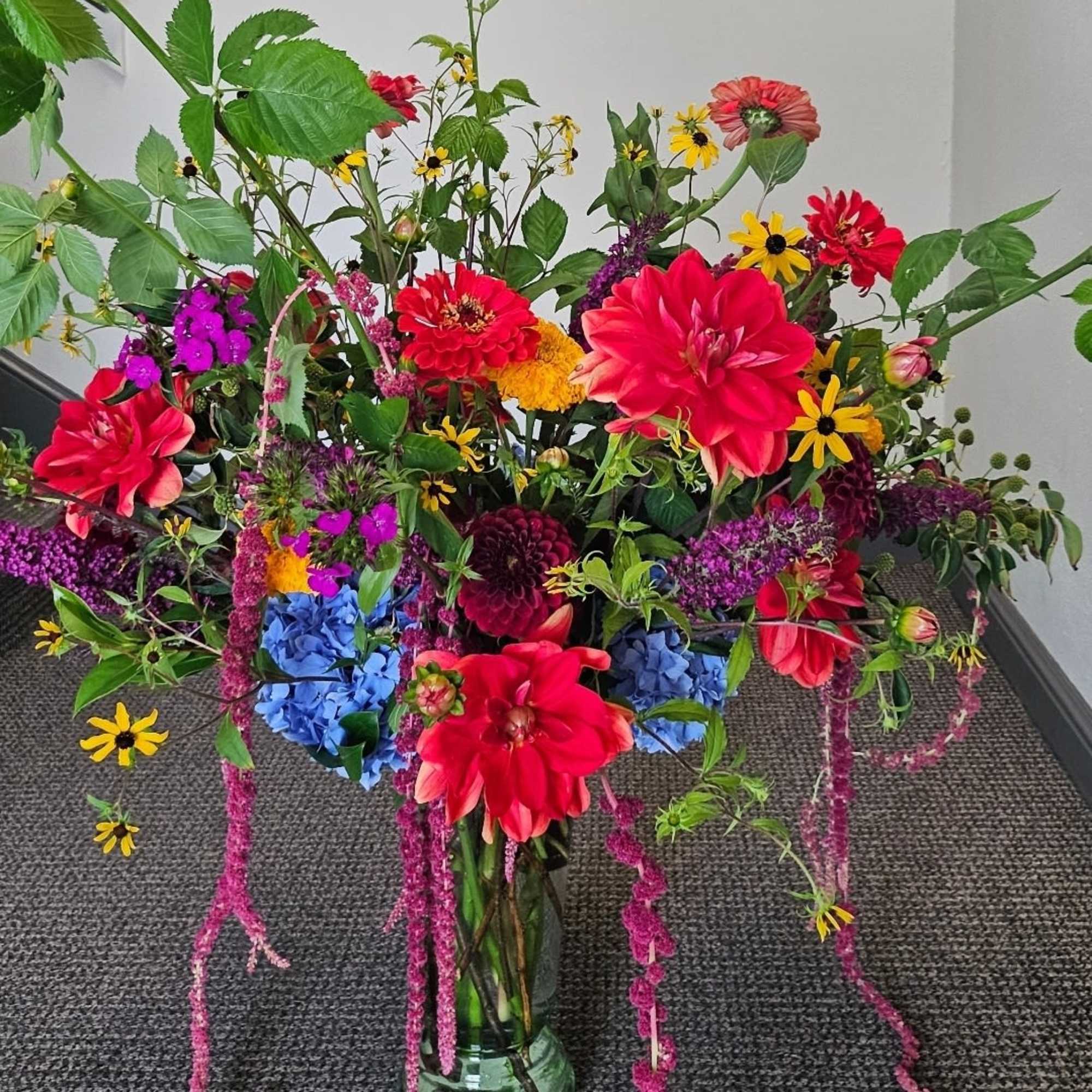 Tall mixed flower arrangement with bright red, yellow, purple, and blue blooms in a clear glass vase