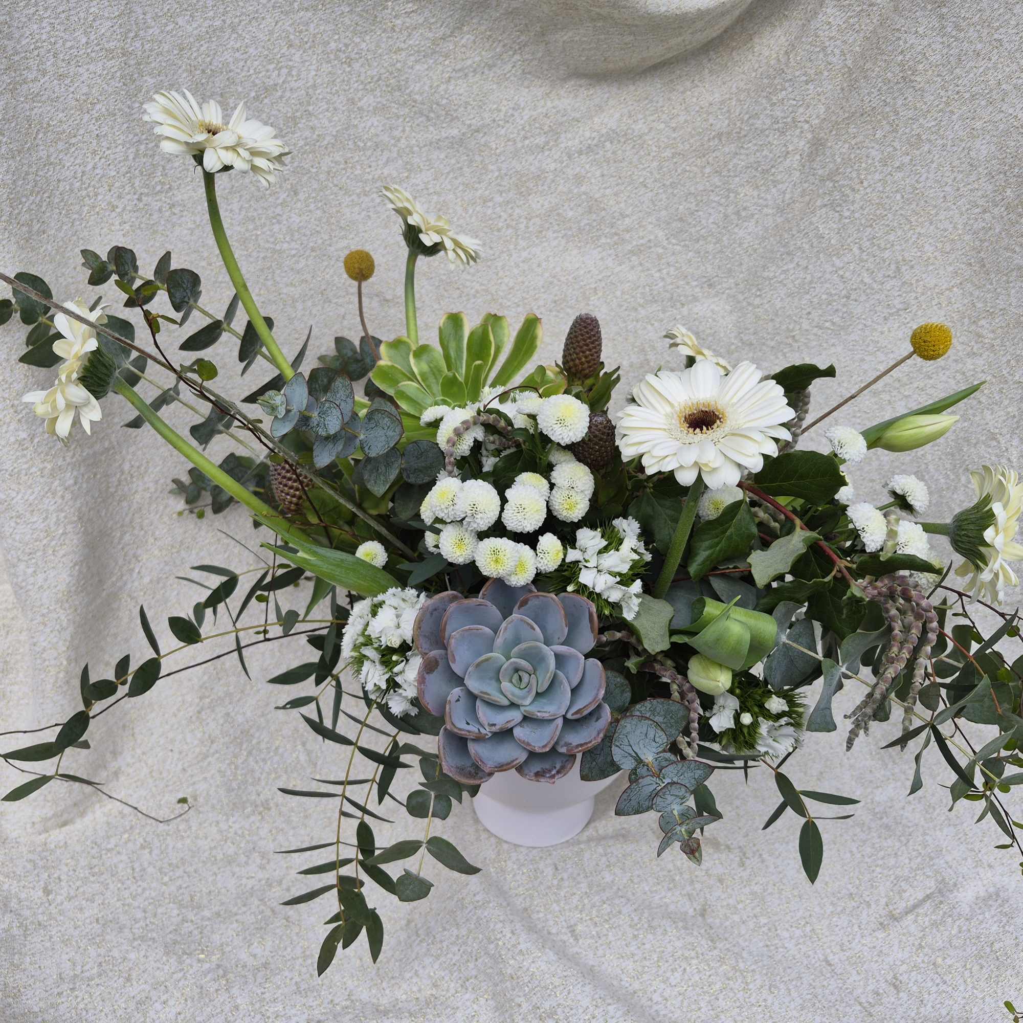 Asymmetrical arrangement with white gerbera daisies, button mums, yellow billy balls, and a central succulent in a white pot