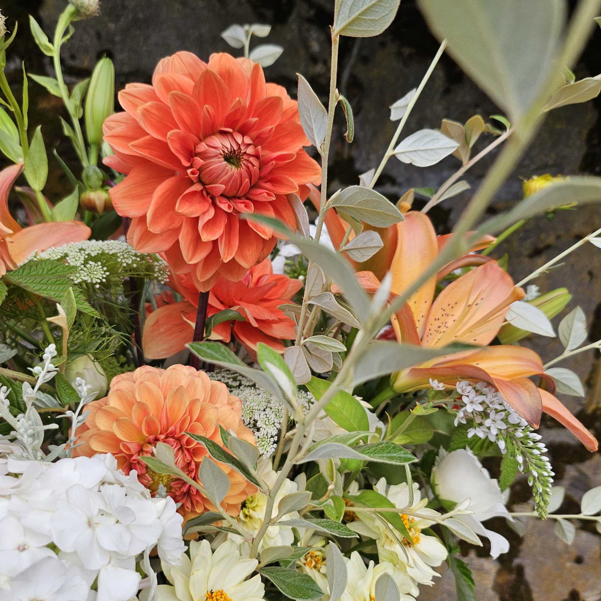 Mixed bouquet with orange dahlias, peach lilies, and white flowers against a dark stone background