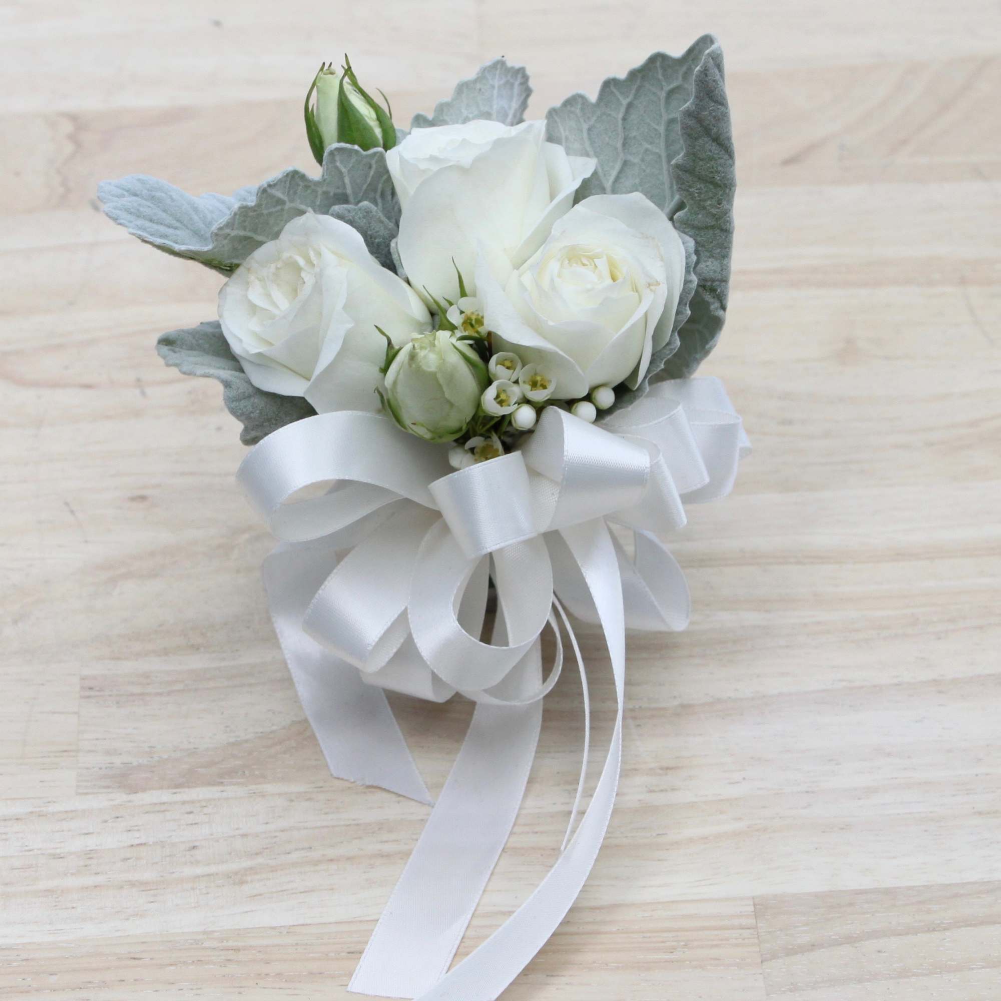 Small white rose corsage with silver foliage and white ribbon bow on a light wood surface