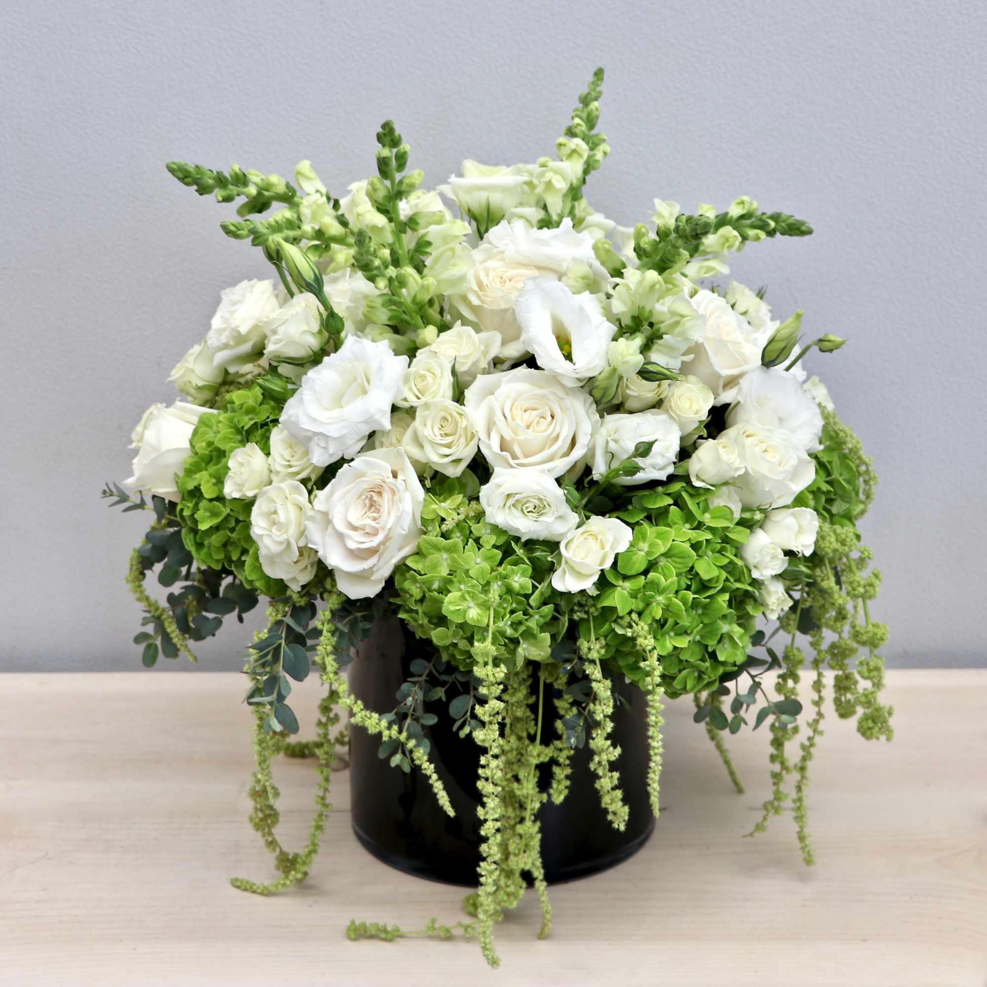 White roses and lisianthus with green hydrangeas in a black cylinder vase with cascading green blooms.