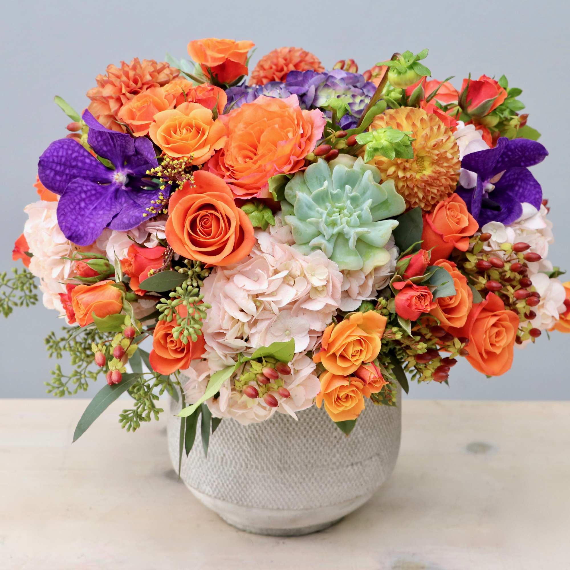 Vibrant arrangement of orange roses, purple orchids, hydrangeas, and a succulent in a white bowl vase