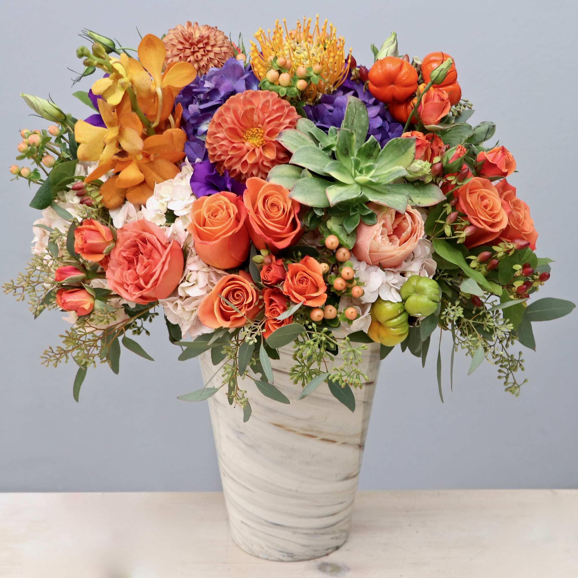 Tall arrangement of orange roses, dahlias, purple blooms and succulent in a white marbled vase