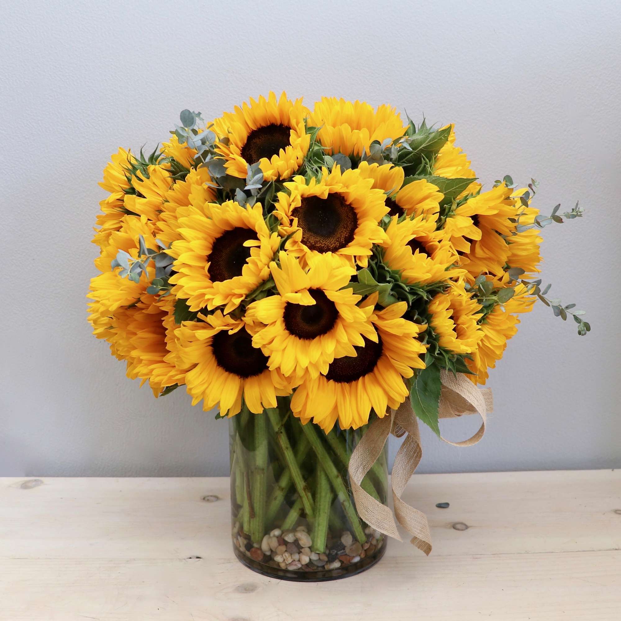 Tall bouquet of yellow sunflowers in a clear glass cylinder vase with pebbles and a burlap ribbon.