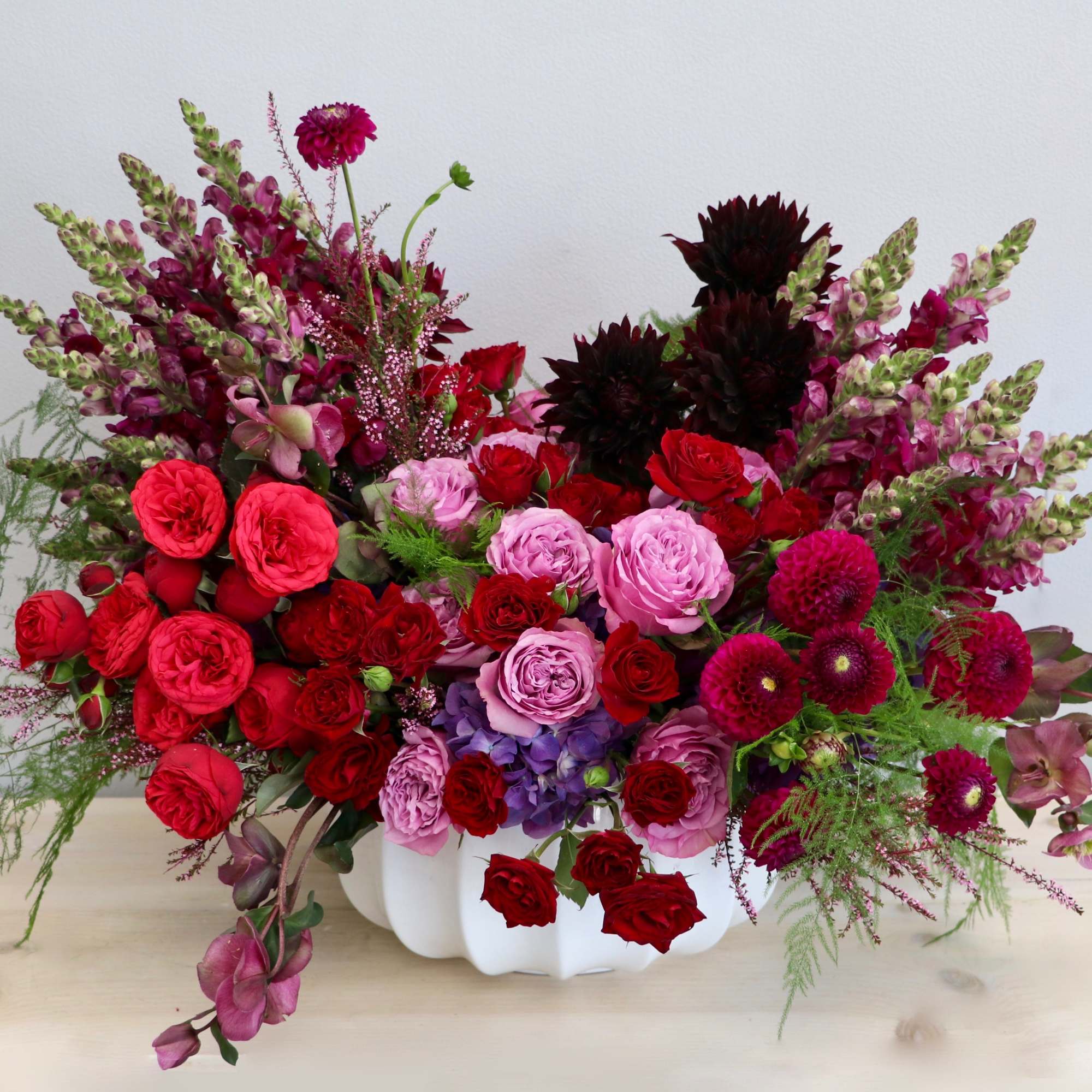 Low white vase filled with rich red, pink, and purple roses, dahlias, and snapdragons in a lush arrangement