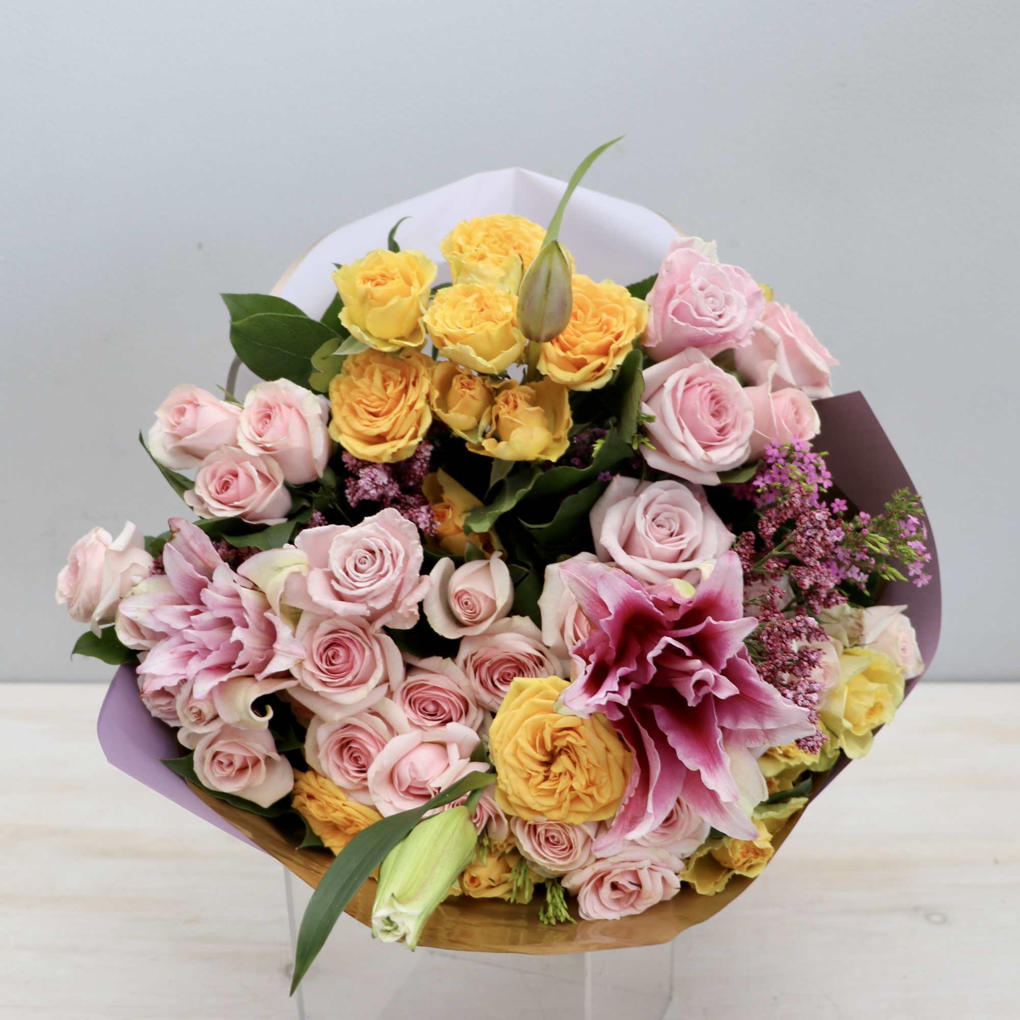 Hand-tied bouquet of pink roses, yellow roses, and pink lilies wrapped in pastel paper.