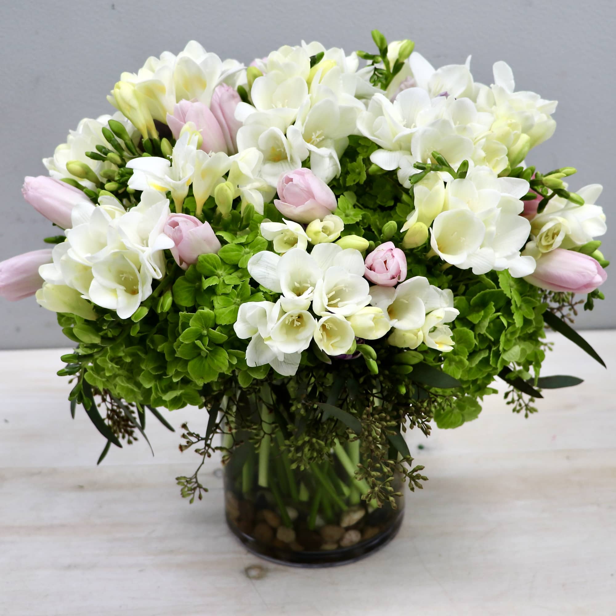 Round arrangement of white freesias, pink tulips, and green hydrangeas in a clear glass vase