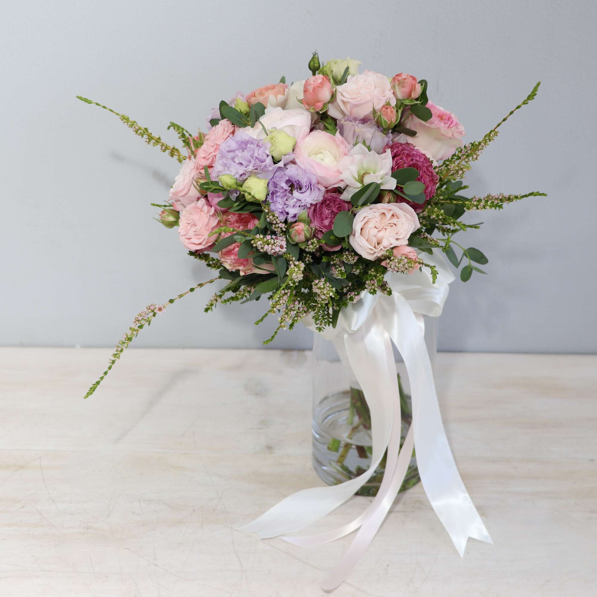 Hand-tied bouquet of pink and white roses with lavender blooms in a glass vase with long white ribbon