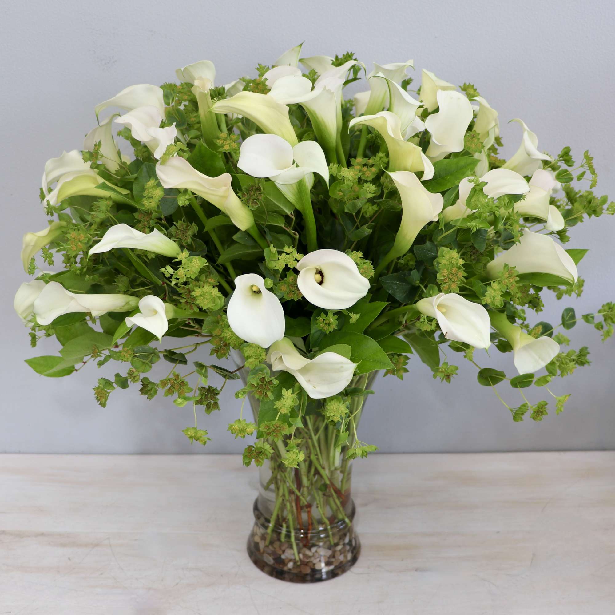 Tall arrangement of white calla lilies with abundant green accents in a clear glass vase
