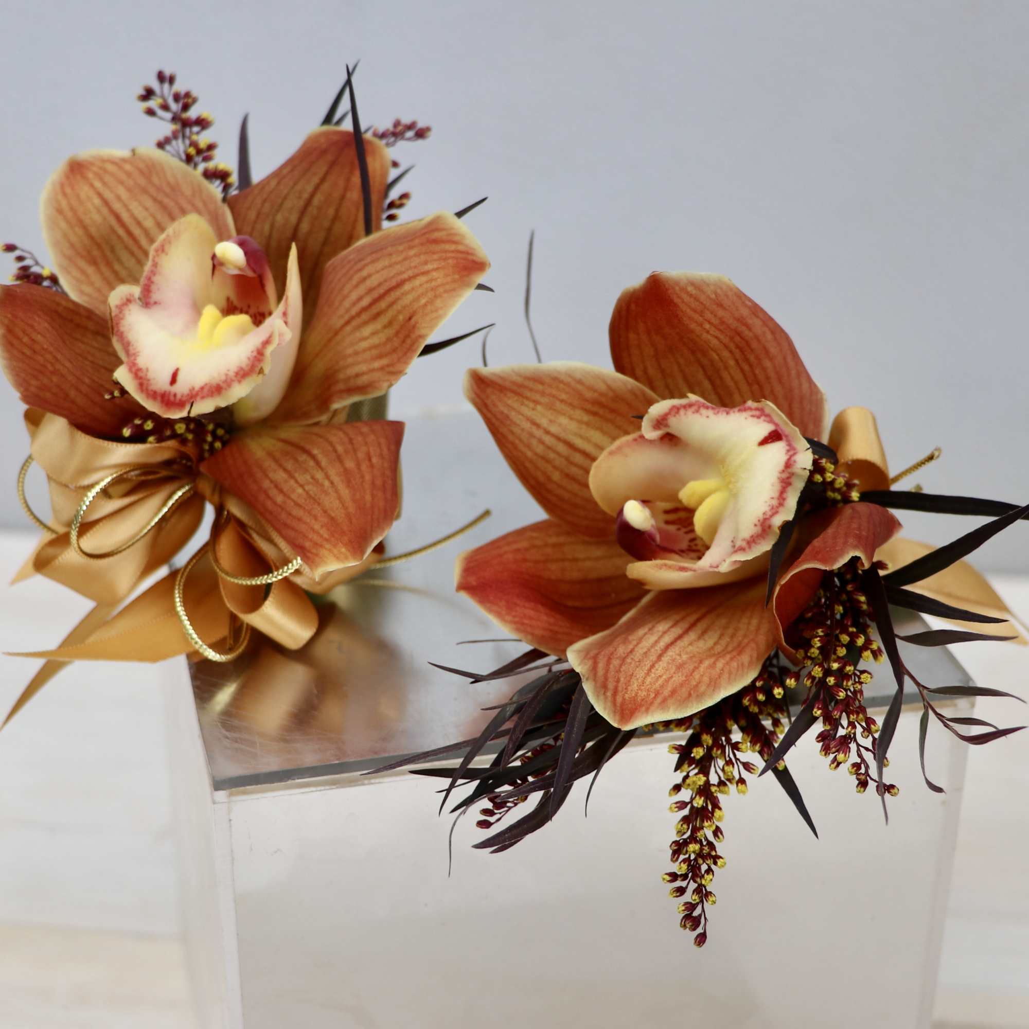Pair of orange cymbidium orchid corsages with gold ribbon and dark accent foliage.