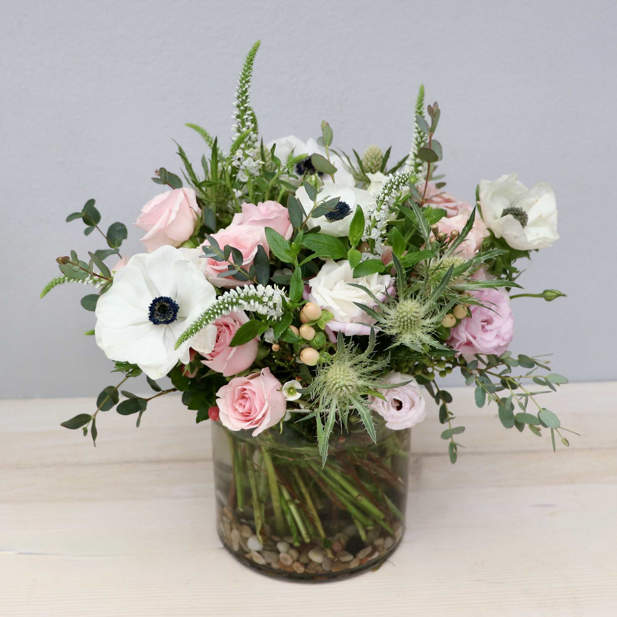 Pastel pink roses and white anemones arranged in a glass cylinder vase with stones.