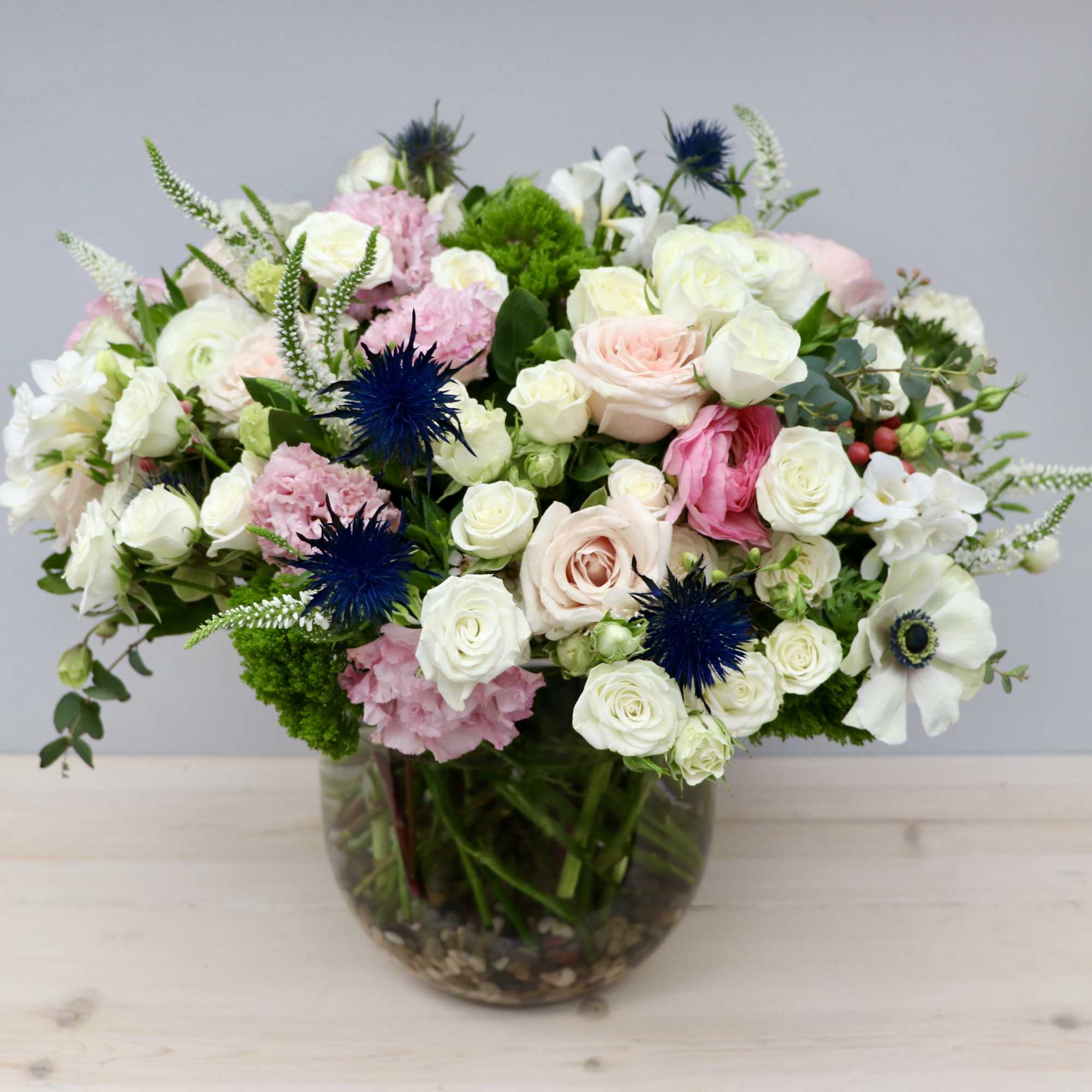 Round arrangement of white and blush roses with blue accents in a clear glass bowl vase