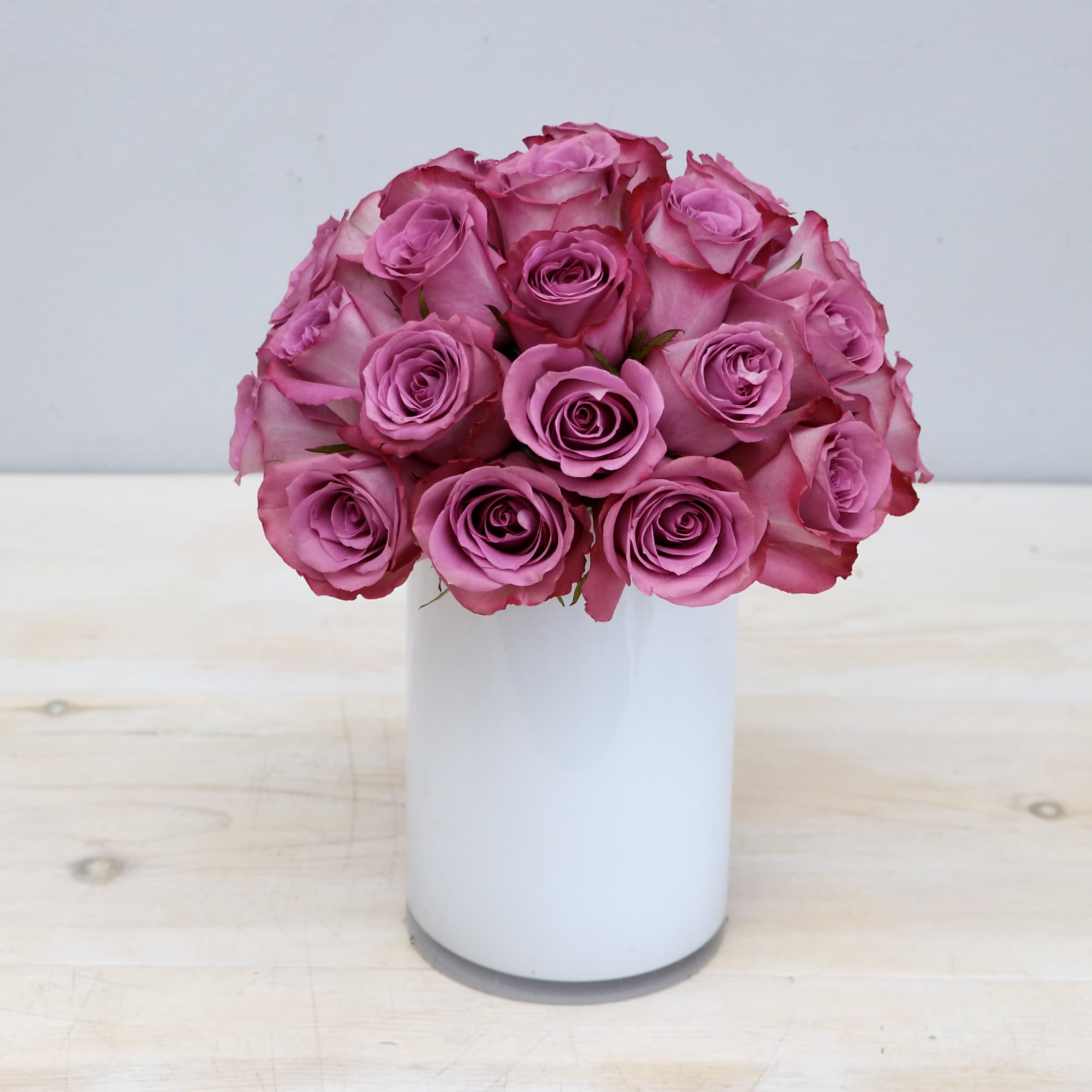 Tight cluster of mauve roses arranged in a white cylindrical vase on a light wood surface