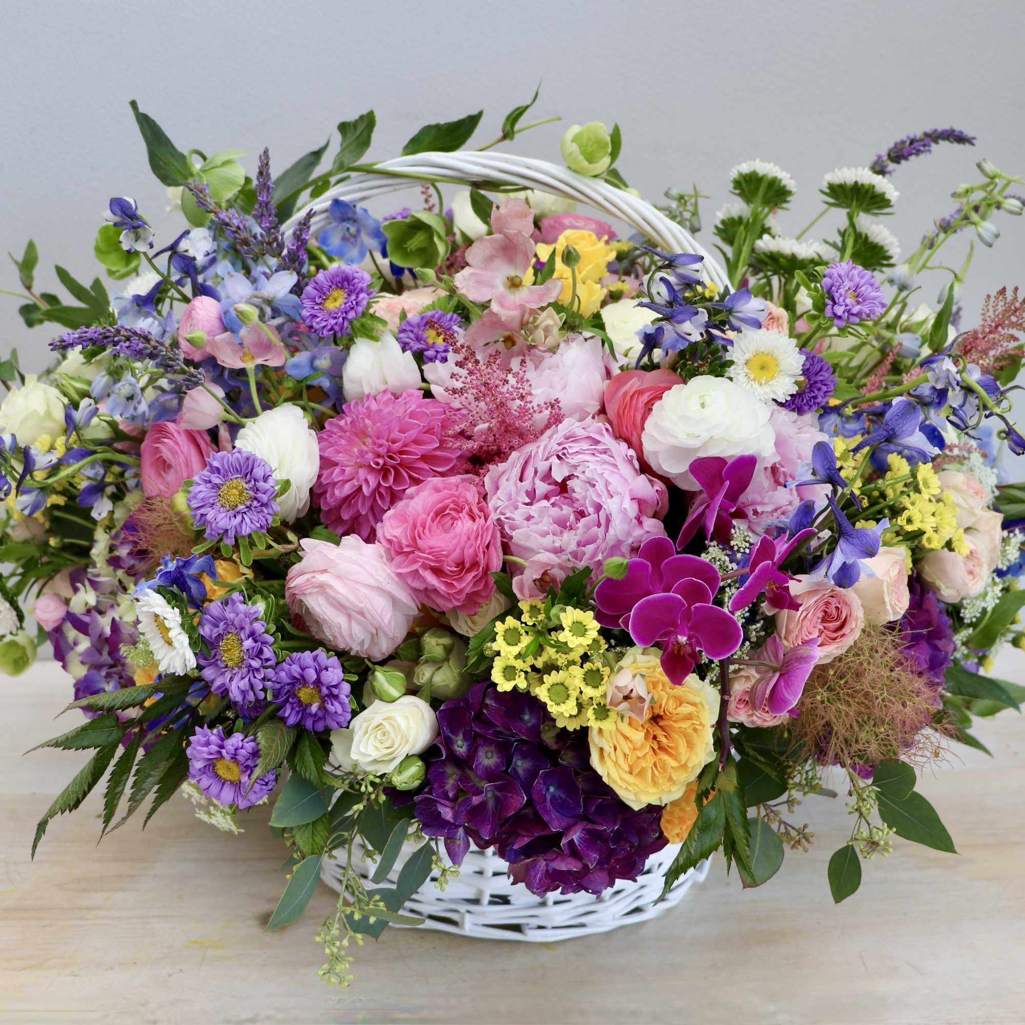 Abundant basket arrangement of pink, purple, white, and yellow mixed flowers in a white wicker basket