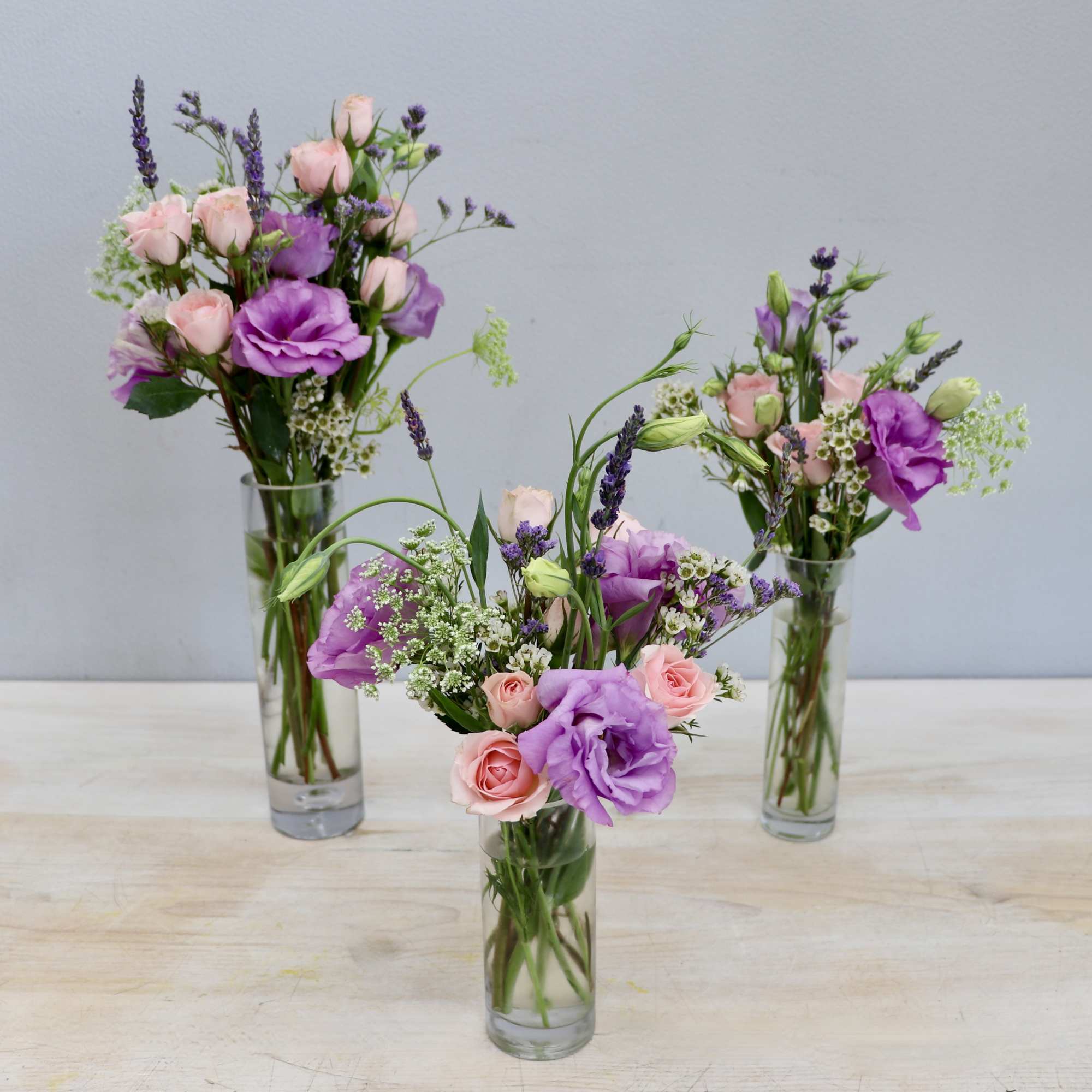 Three slim glass vases with pink spray roses and purple lisianthus in loose arrangements