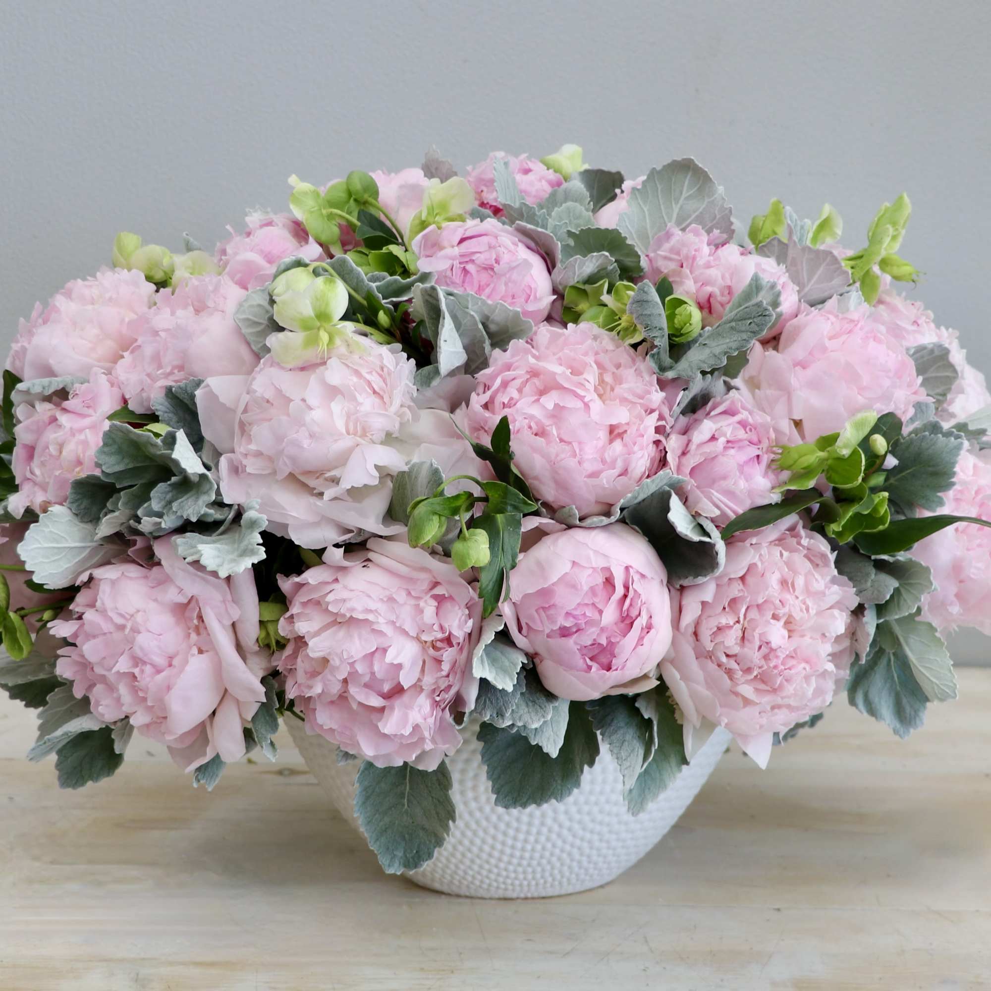 Low white bowl filled with lush light pink peonies and soft gray foliage.