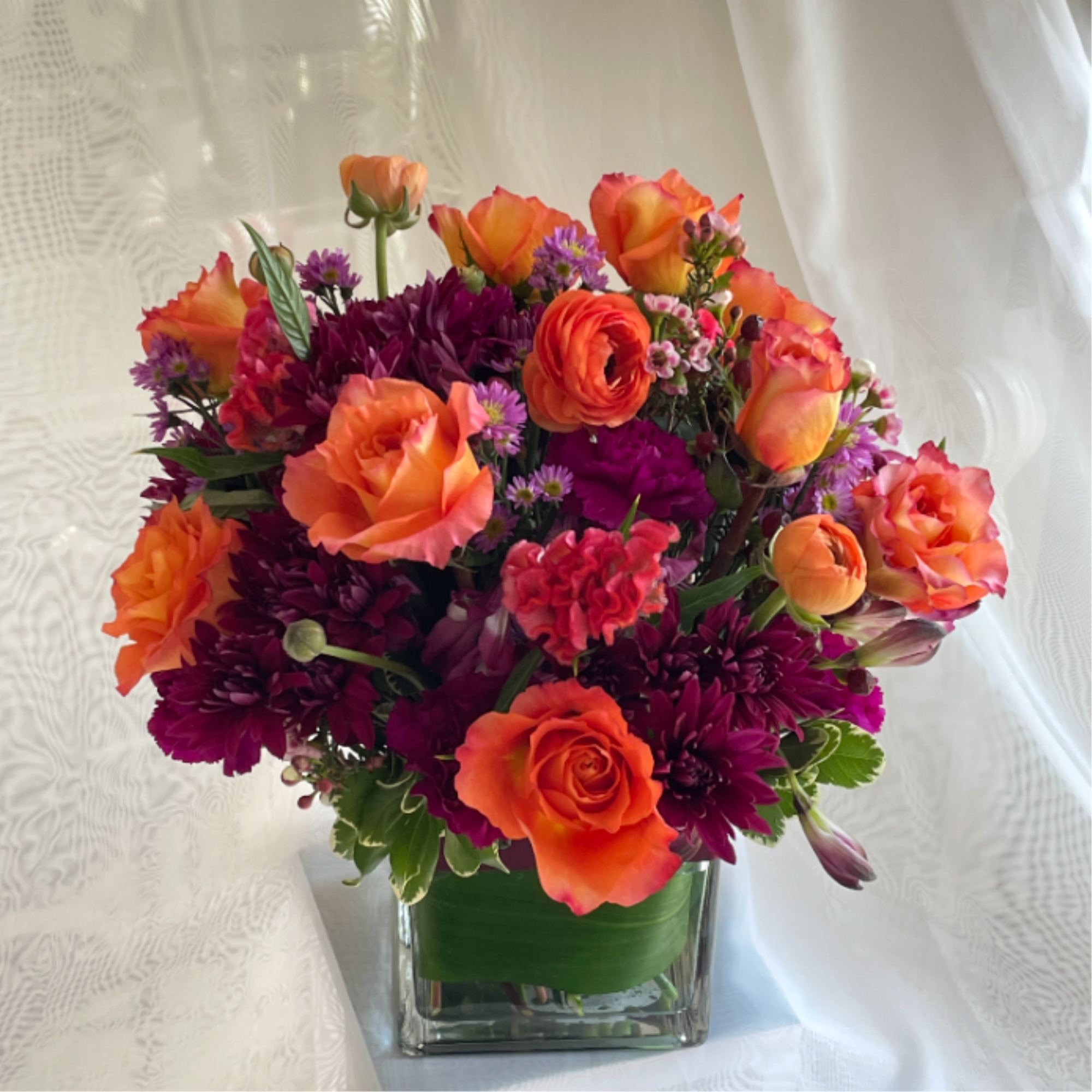 Orange roses and purple flowers arranged in a square glass vase