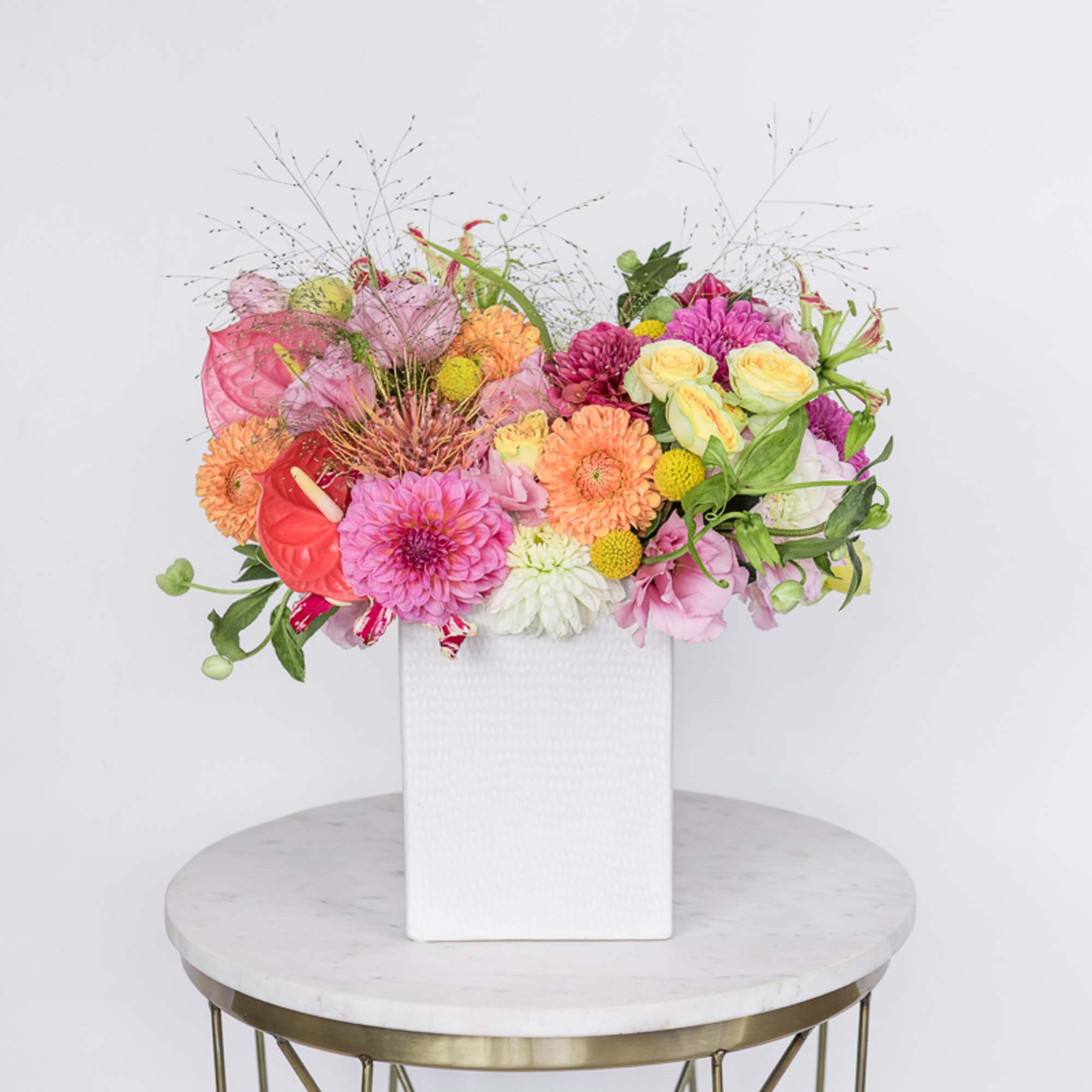 Bright mixed arrangement of pink, orange, and yellow flowers in a white rectangular vase on a round table.