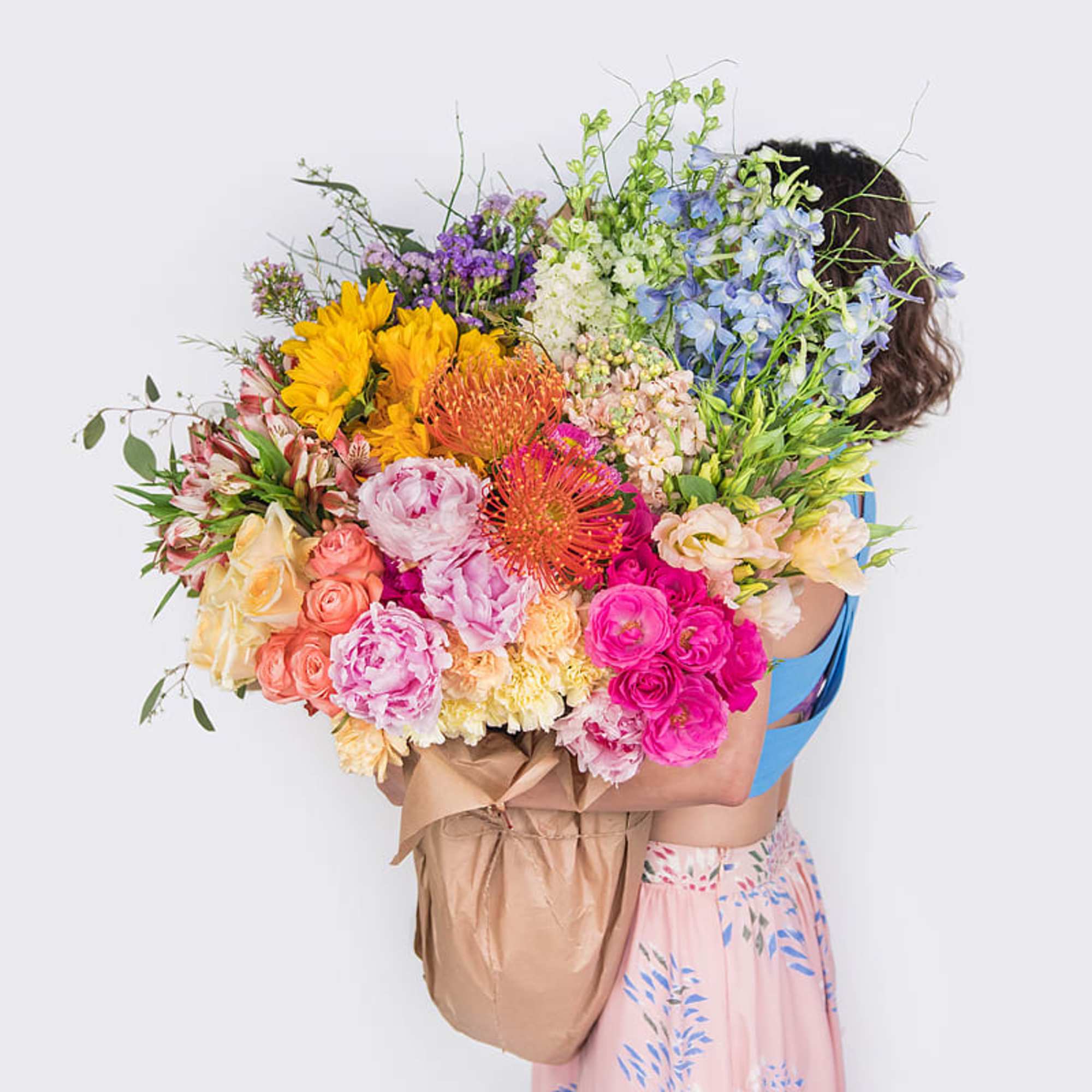 Person holding an oversized mixed bouquet of bright roses, sunflowers, peonies, and other flowers wrapped in kraft paper.