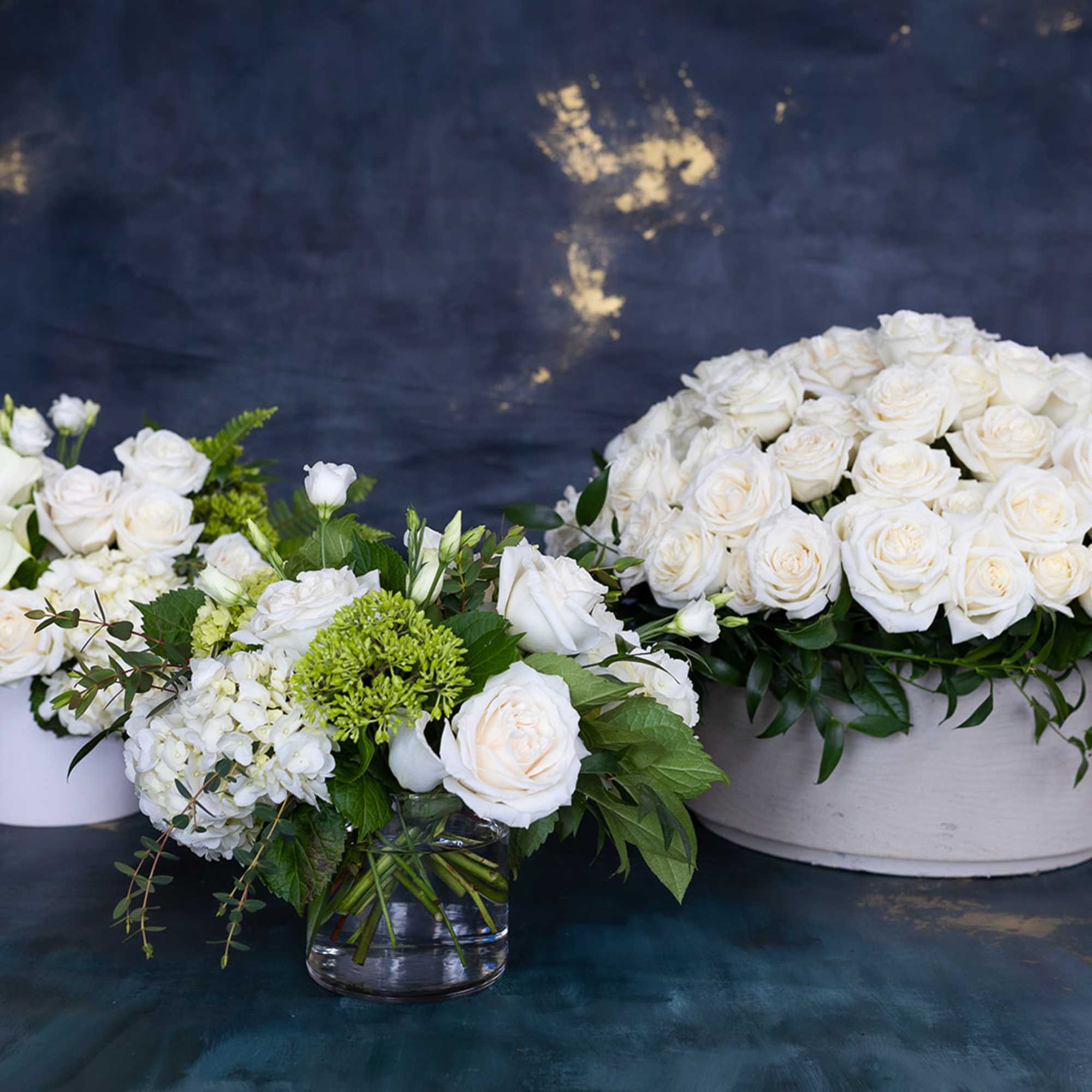 Three white floral arrangements with roses and greenery in white containers