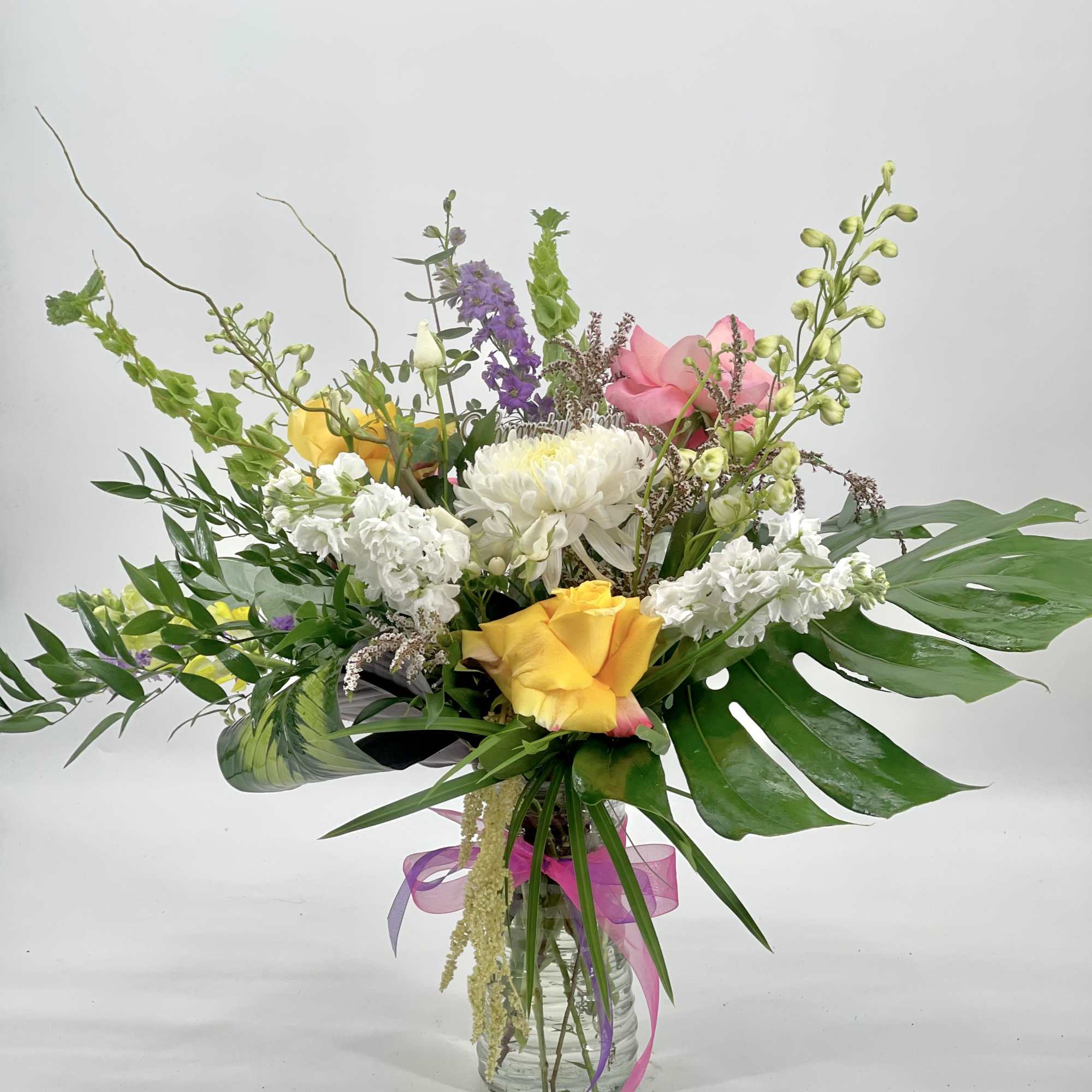 Tall mixed arrangement with yellow and pink roses, white blooms, and large tropical leaves in a clear glass vase