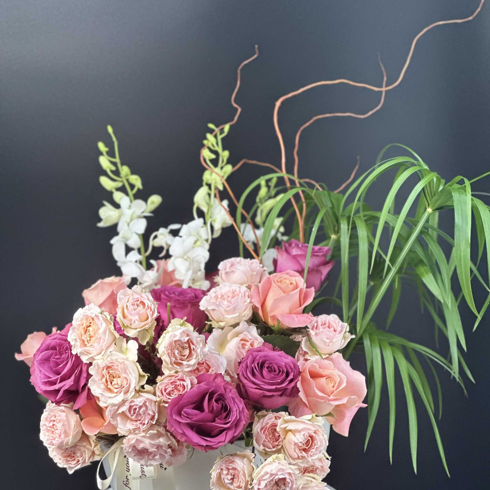 Pink and purple roses with white orchids and palm fronds in a white hatbox arrangement
