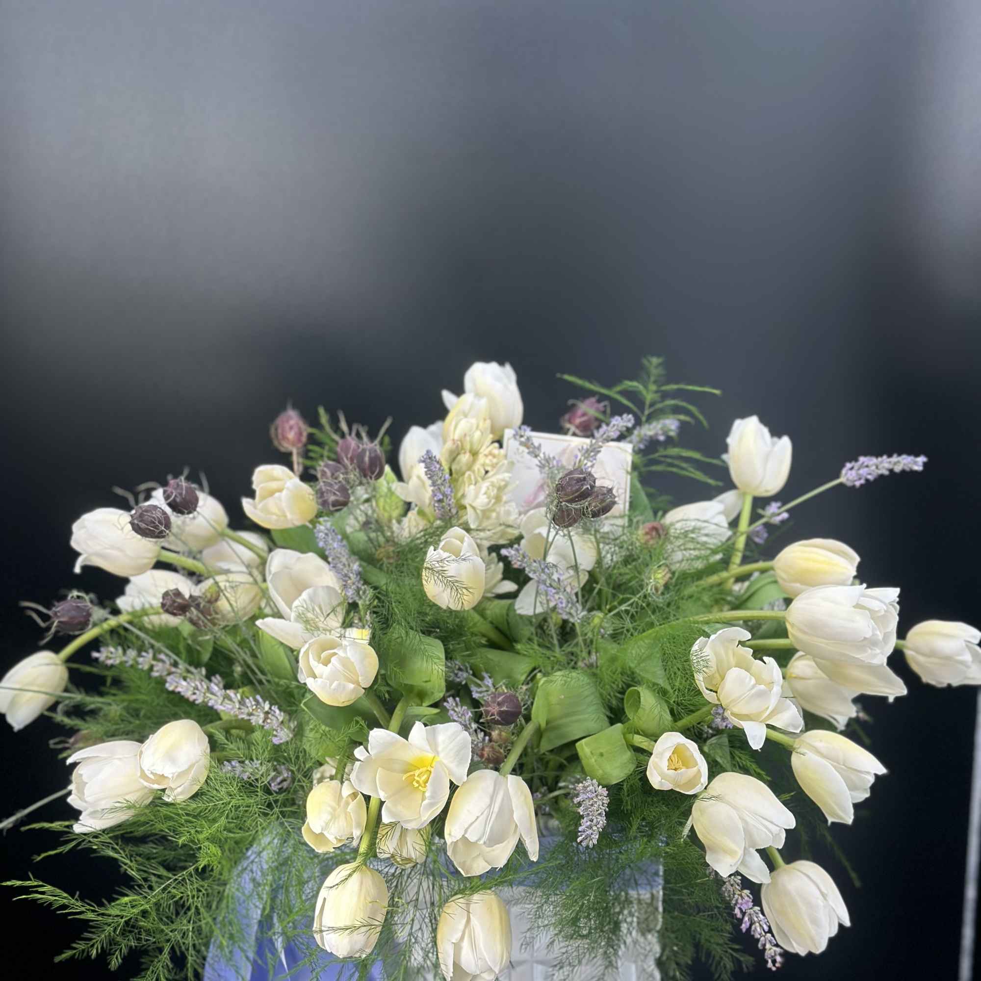 White tulips and lavender filler flowers in a white vase with a blue ribbon