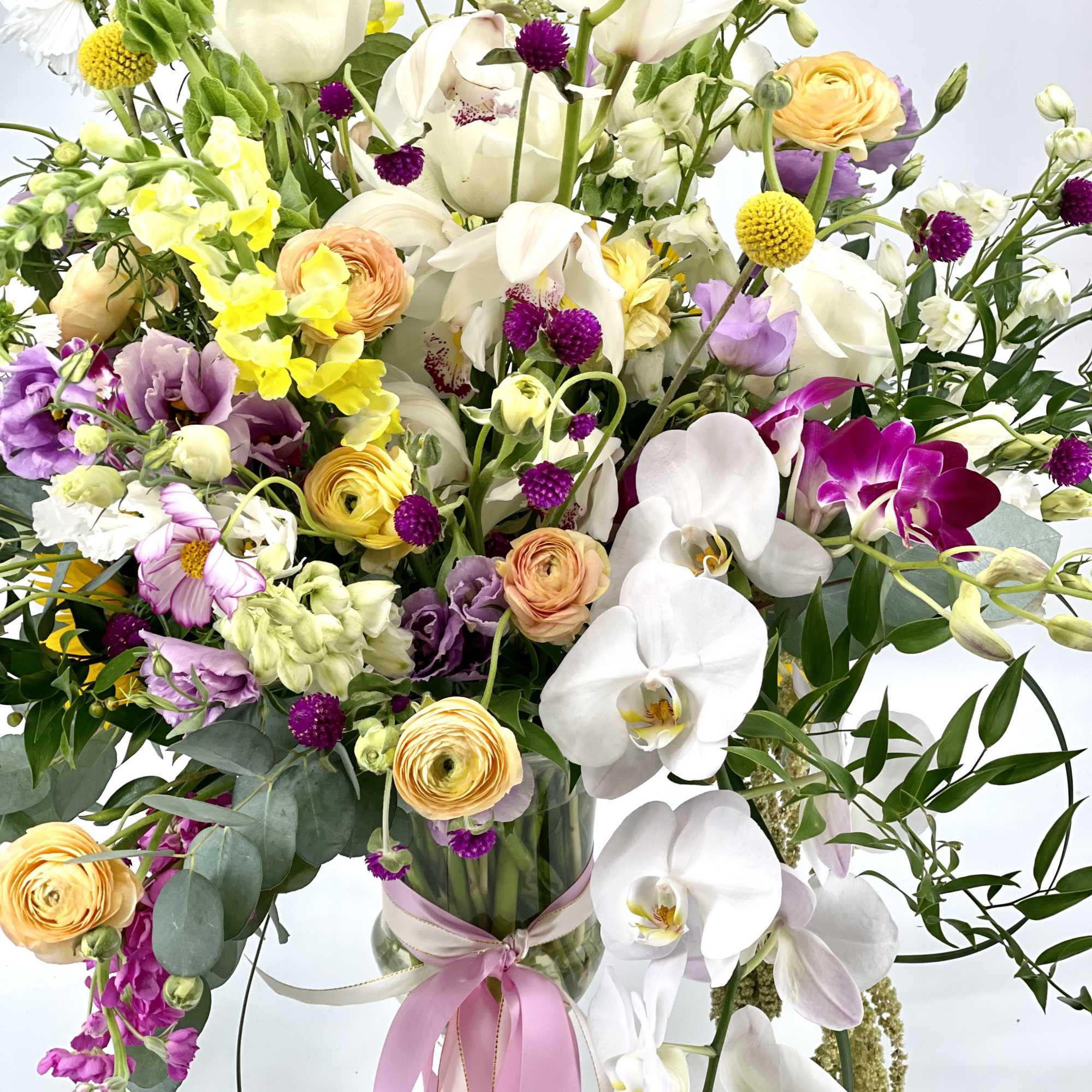 Mixed bouquet of orchids, roses, and ranunculus in a glass vase with a pink ribbon