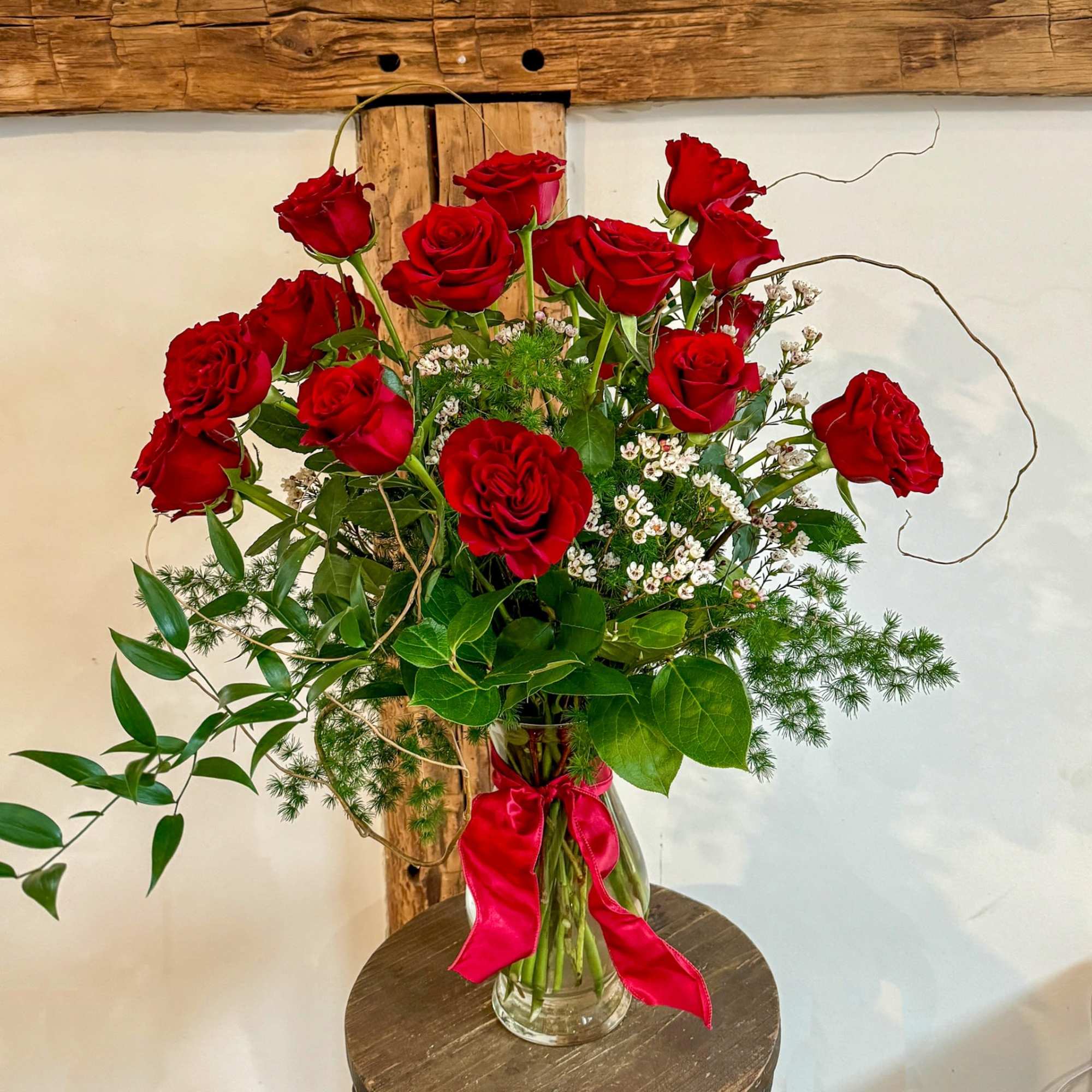 Tall arrangement of red roses with small white blooms in a clear glass vase with a red ribbon.