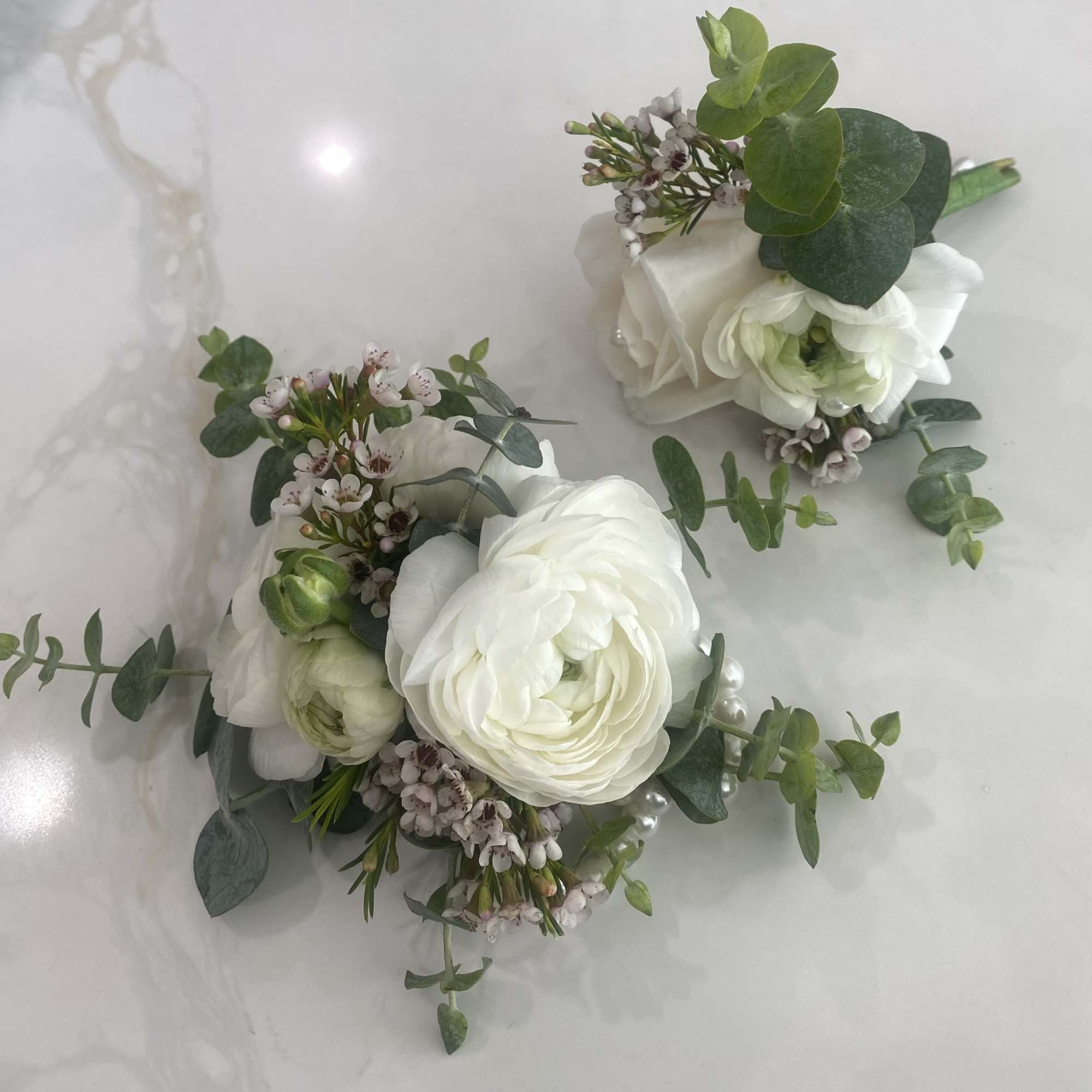 Two white floral boutonnieres with small pink filler flowers and greenery