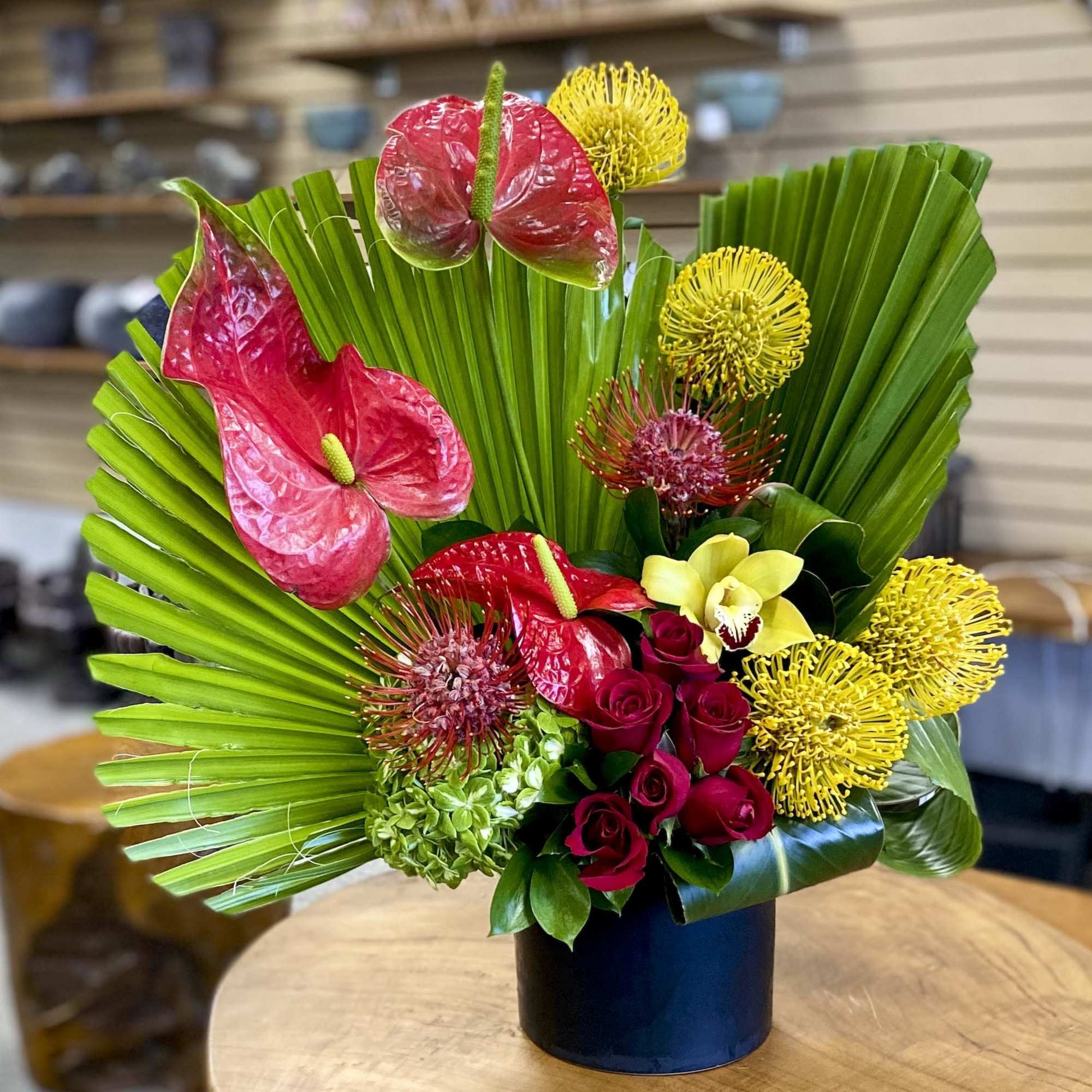 Tropical floral arrangement with red anthuriums, yellow blooms, and roses in a black vase