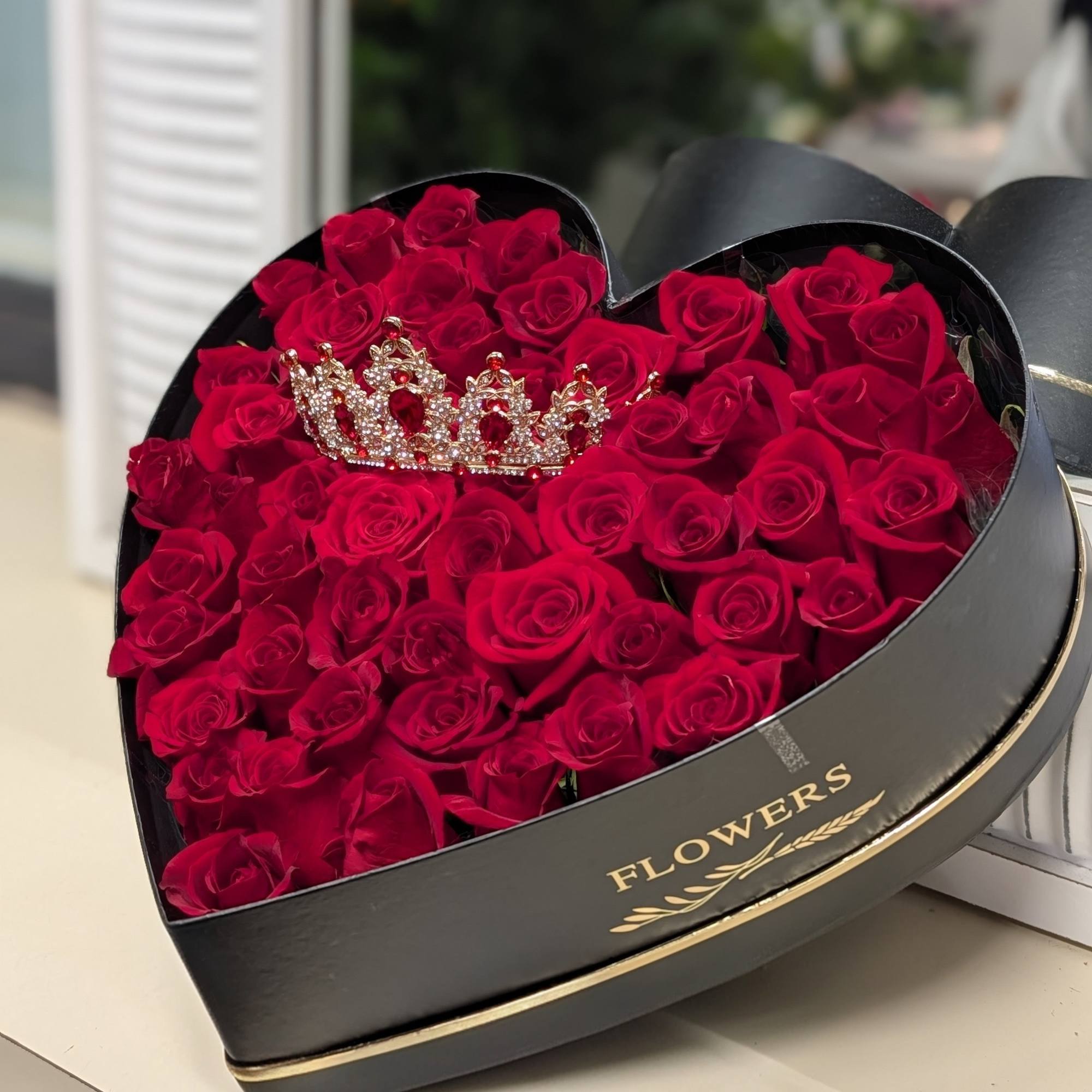 Heart-shaped black box filled with red roses and topped with a jeweled crown ornament.