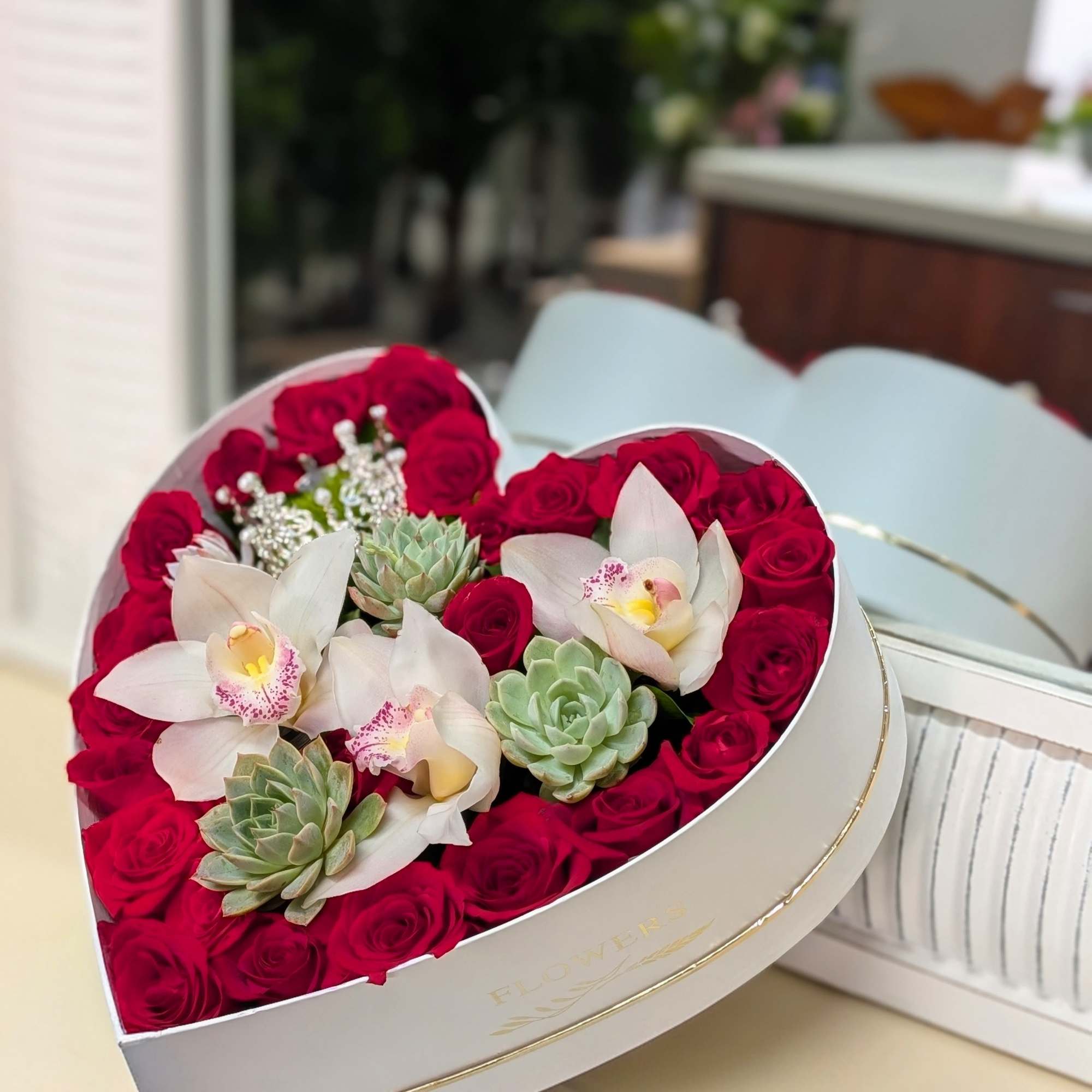 Heart-shaped white box filled with red roses, pale orchids, and green succulents.