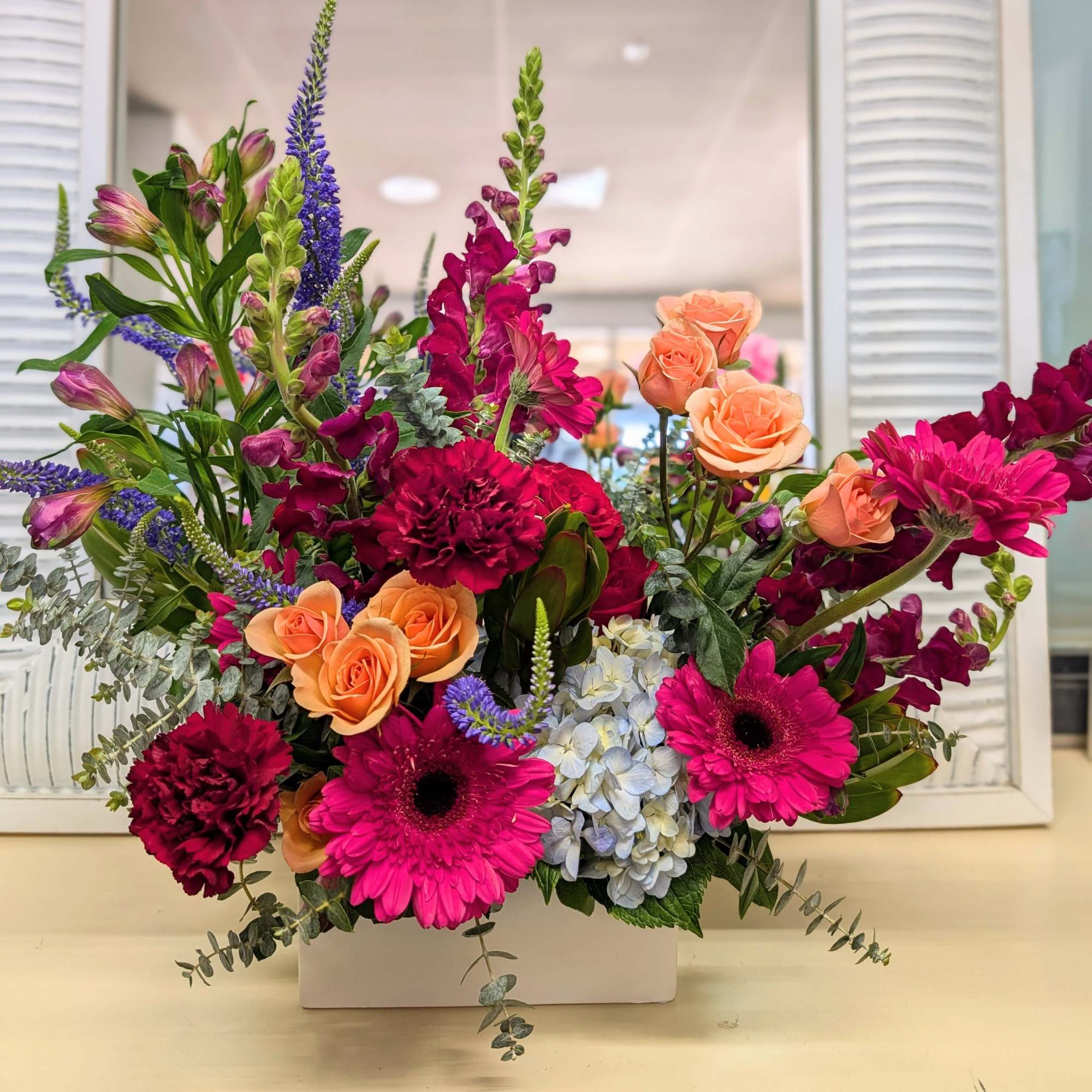 Bright arrangement of hot pink gerberas, peach roses, carnations and hydrangea in a white rectangular vase