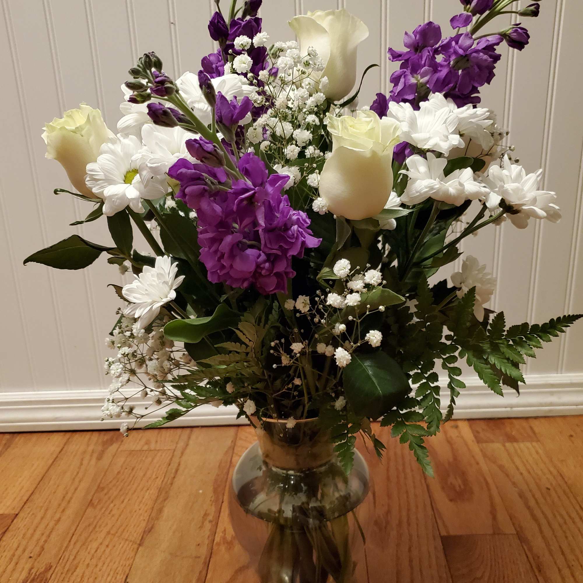 Arrangement of white roses, white daisies, and purple flowers in a clear glass vase on a wood floor