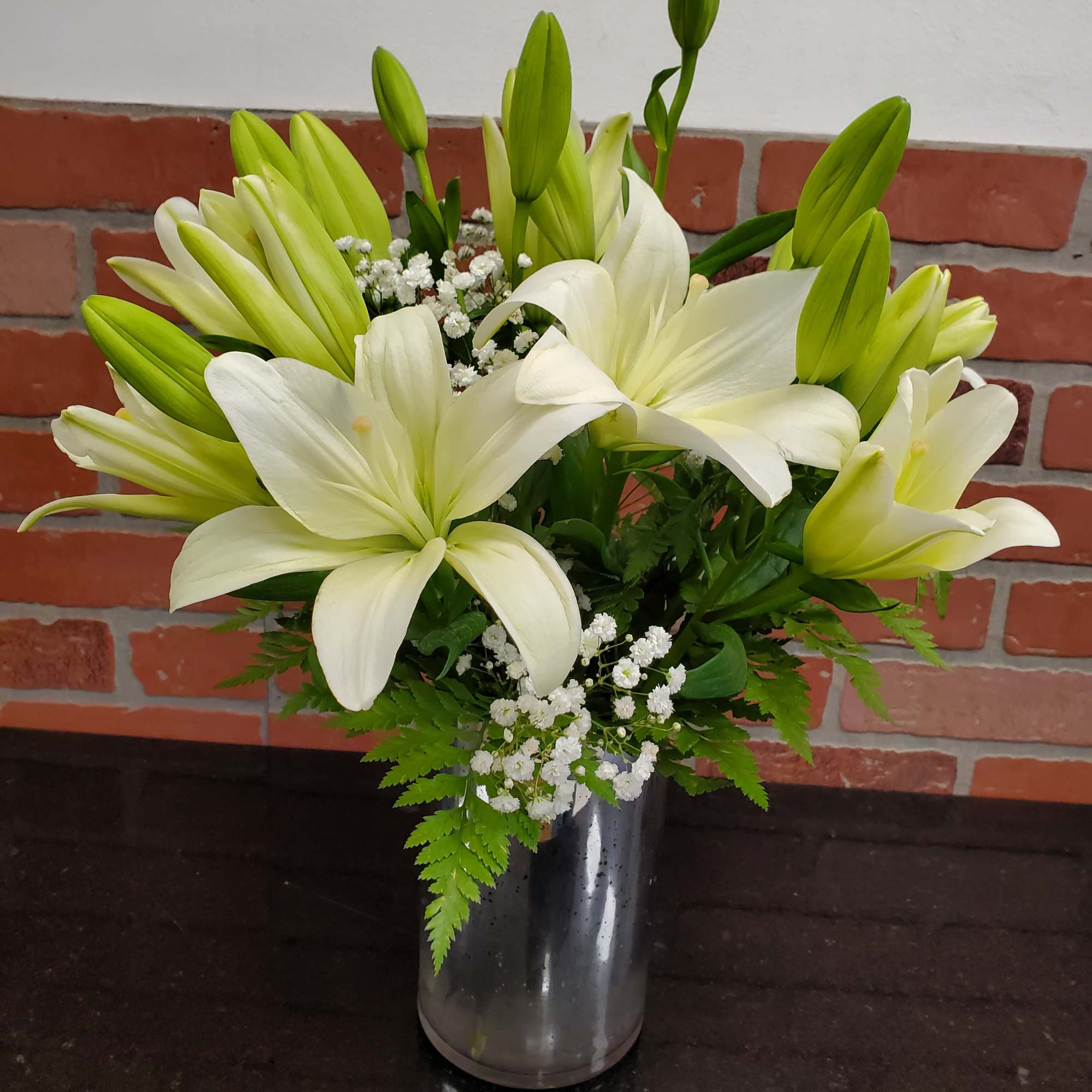 Tall arrangement of white lilies with buds and small white blooms in a glass cylinder vase