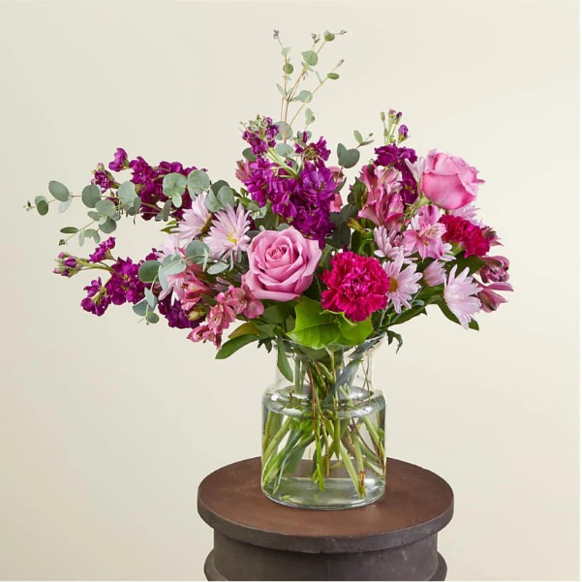 Pink and purple mixed flower arrangement with roses and daisies in a clear glass vase on a small round table