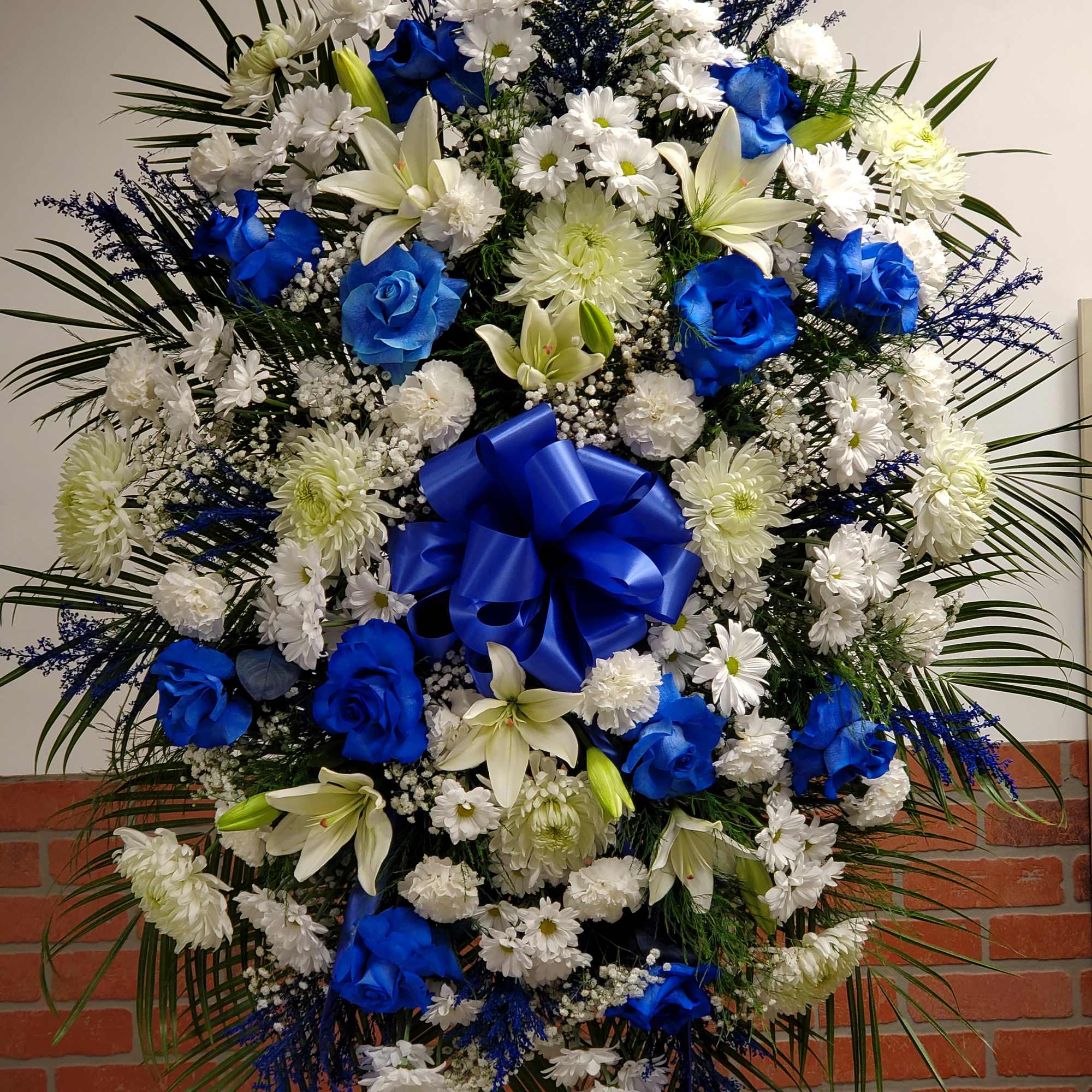 Tall standing spray of blue roses, white lilies, and mixed white flowers with a large blue bow