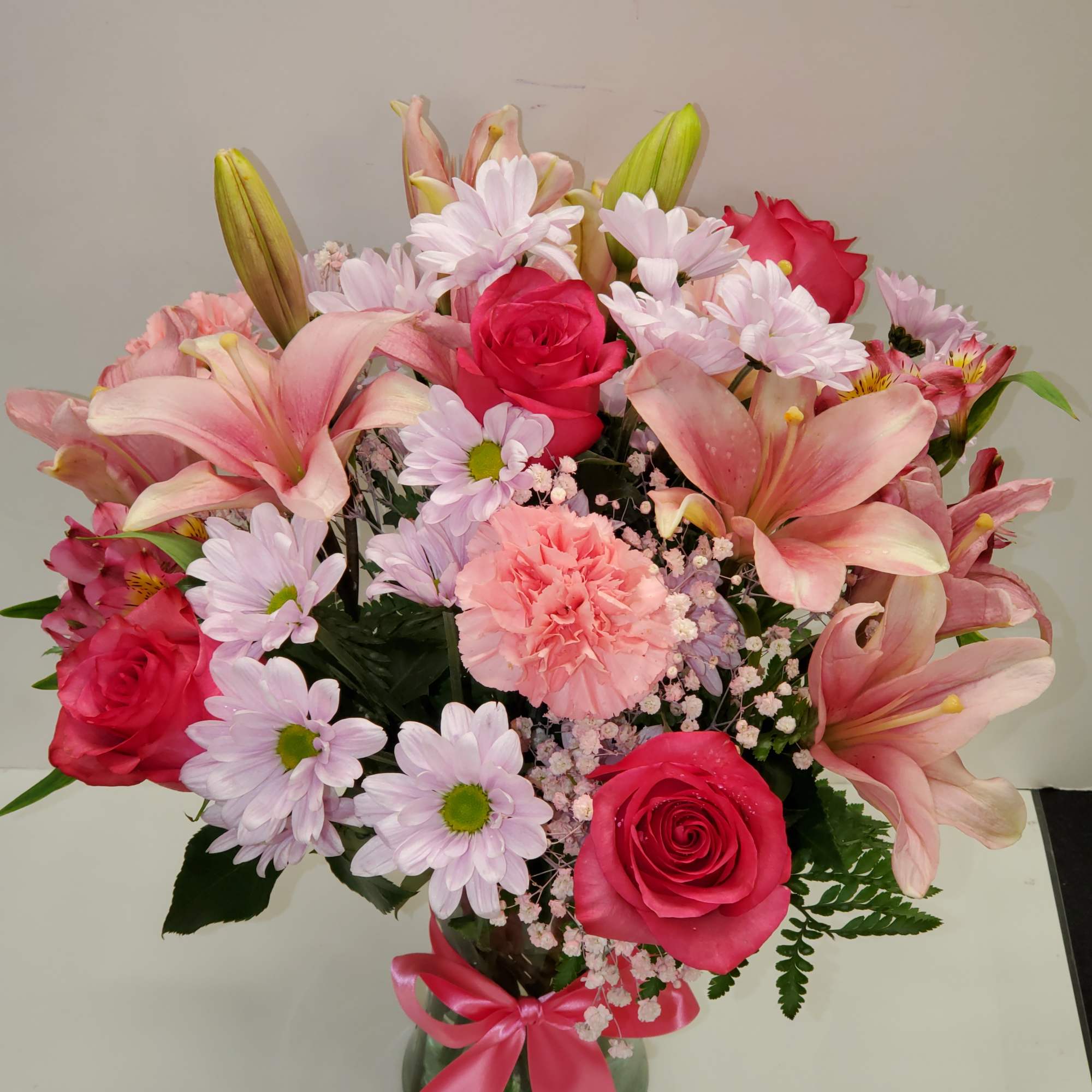 Pink rose, lily, daisy, and carnation arrangement in a clear glass vase with a pink ribbon