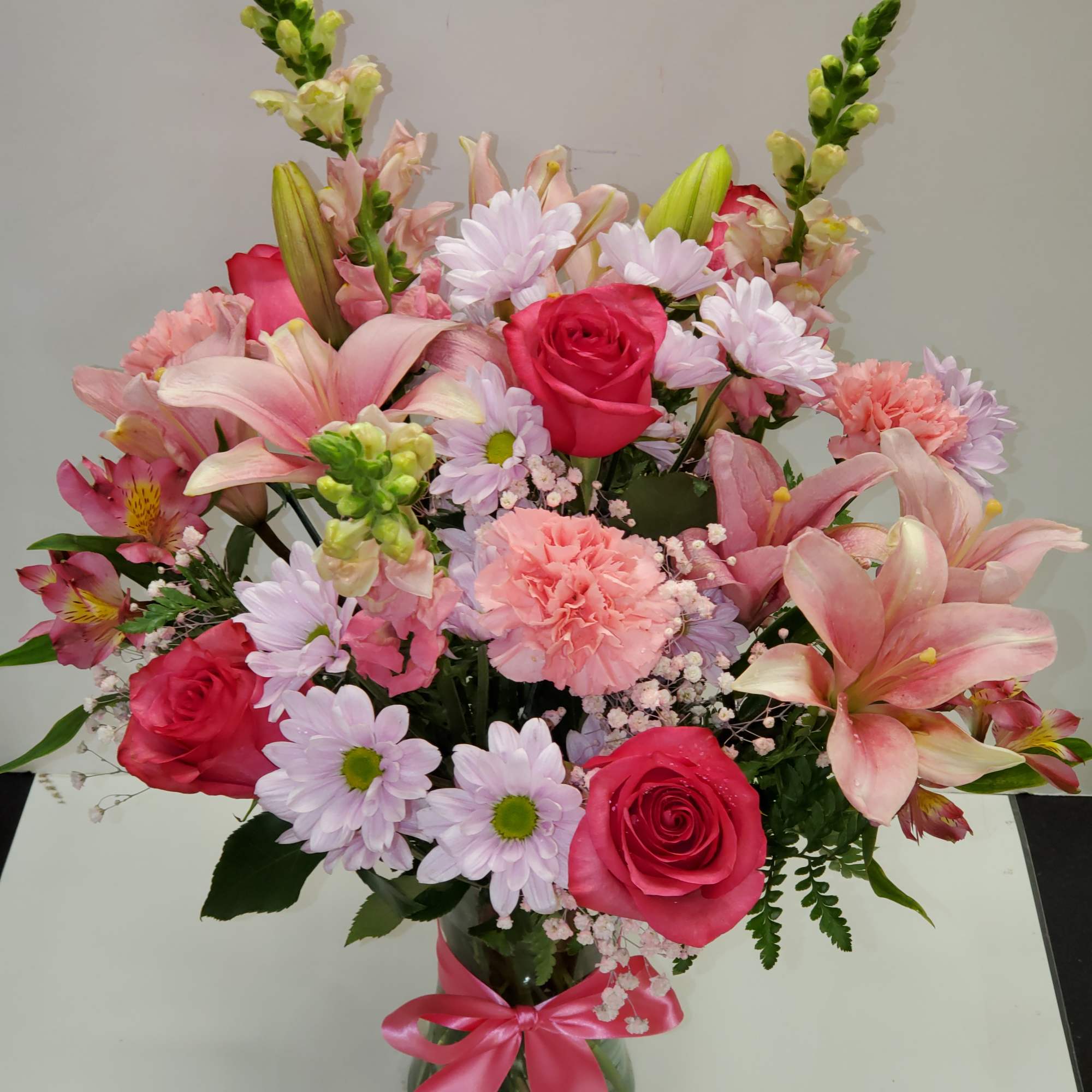 Mixed bouquet of pink roses, lilies, carnations, and daisies in a clear glass vase with a pink ribbon.