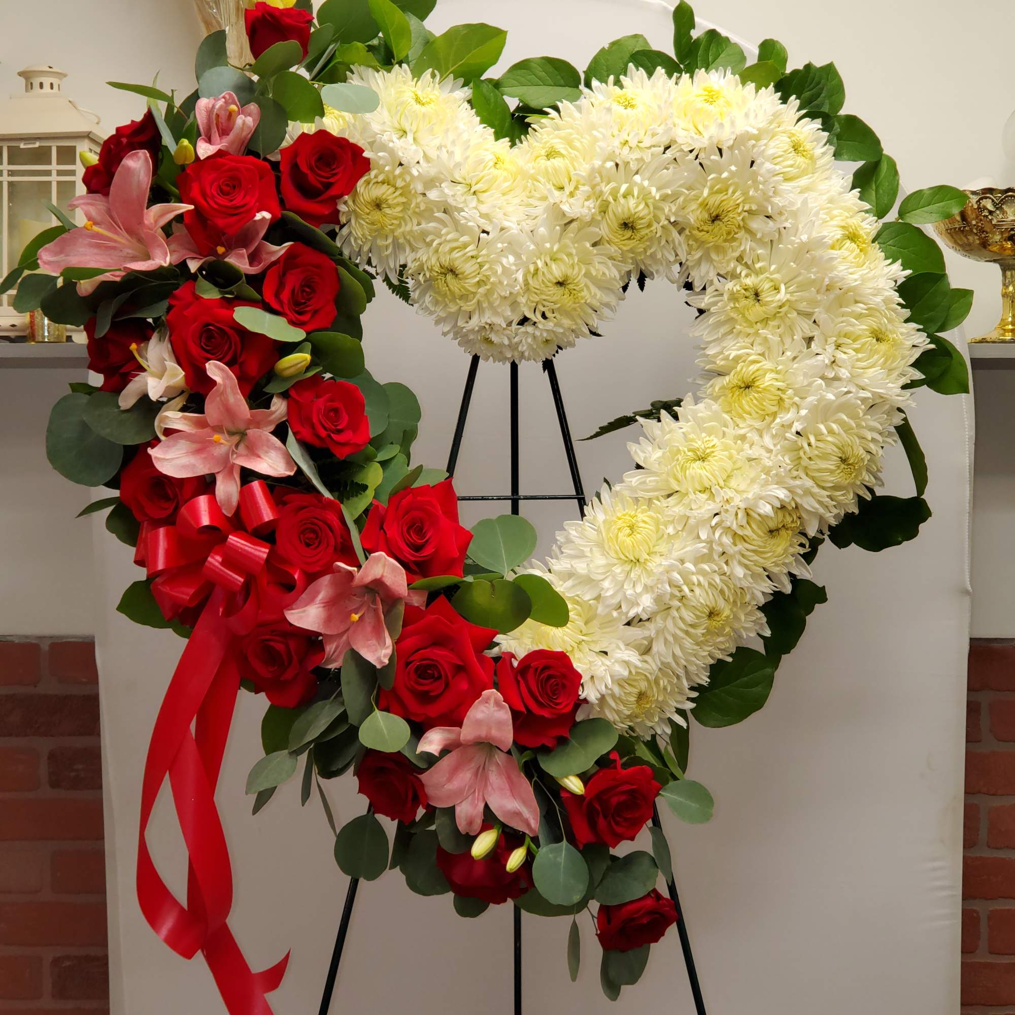 Standing heart-shaped wreath of white mums with red roses and pink lilies on a black easel